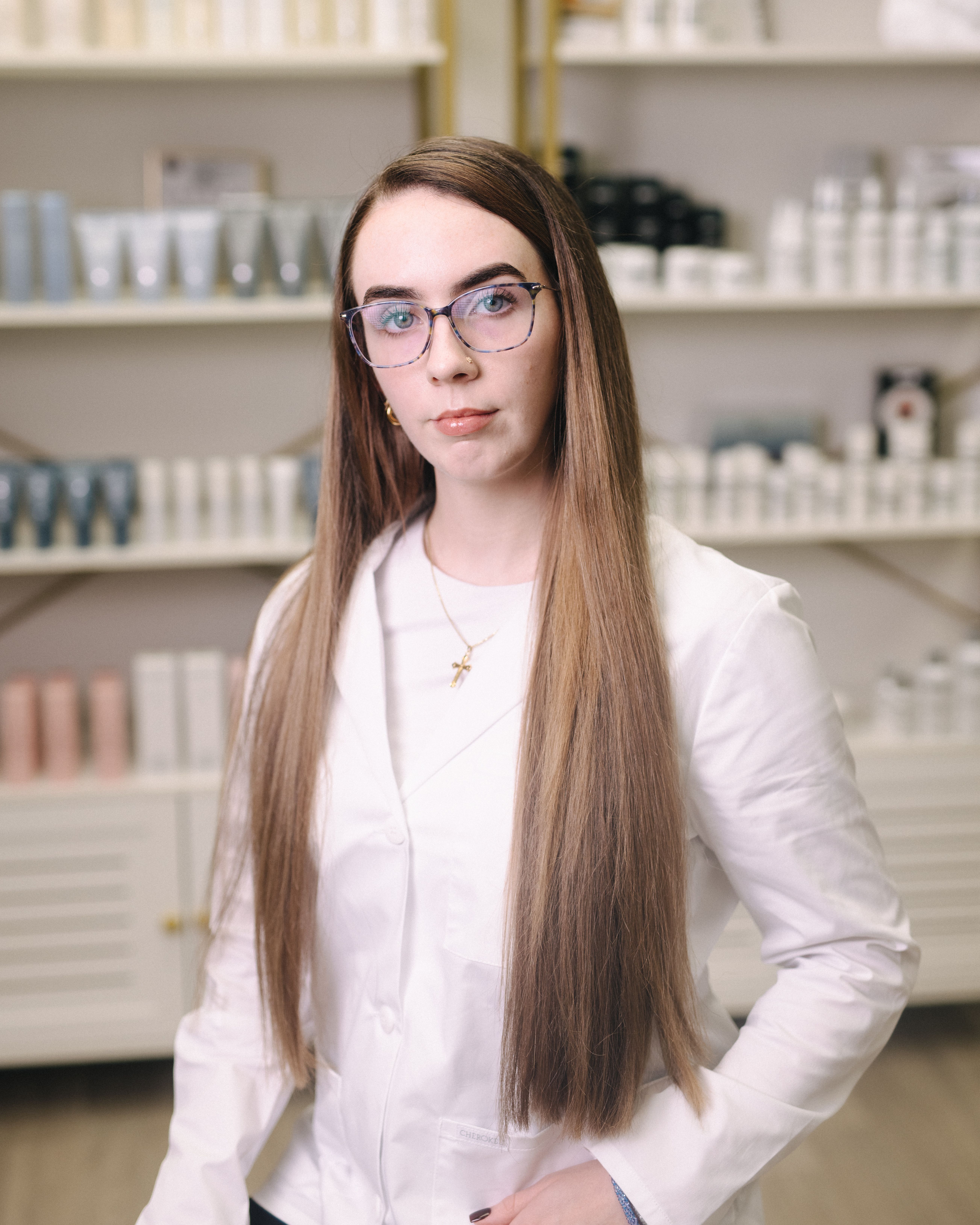 A woman with long brown hair, glasses, and a white lab coat stands in front of product shelves.