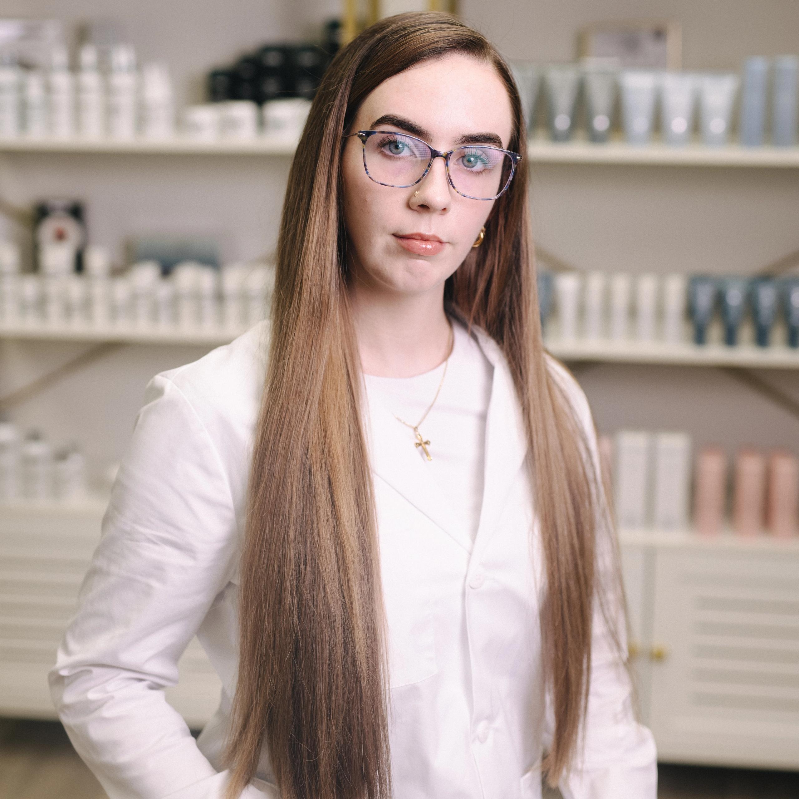 A woman with long brown hair, glasses, and a white lab coat stands in front of product shelves.