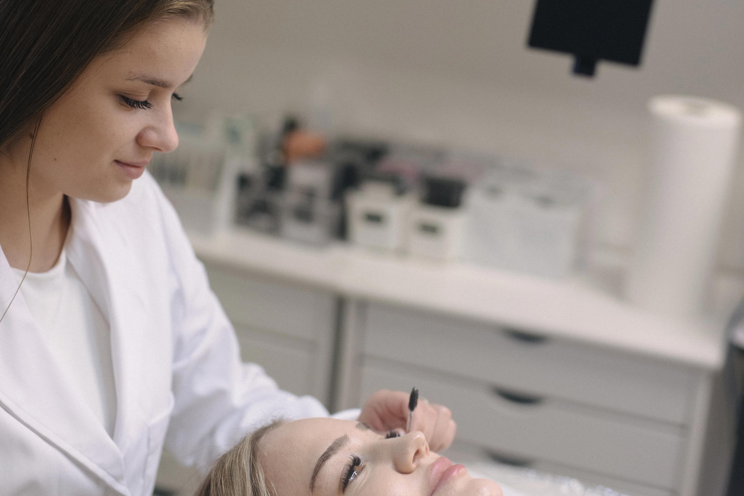 A beauty professional in a white coat applies a product to a client's eyelashes.