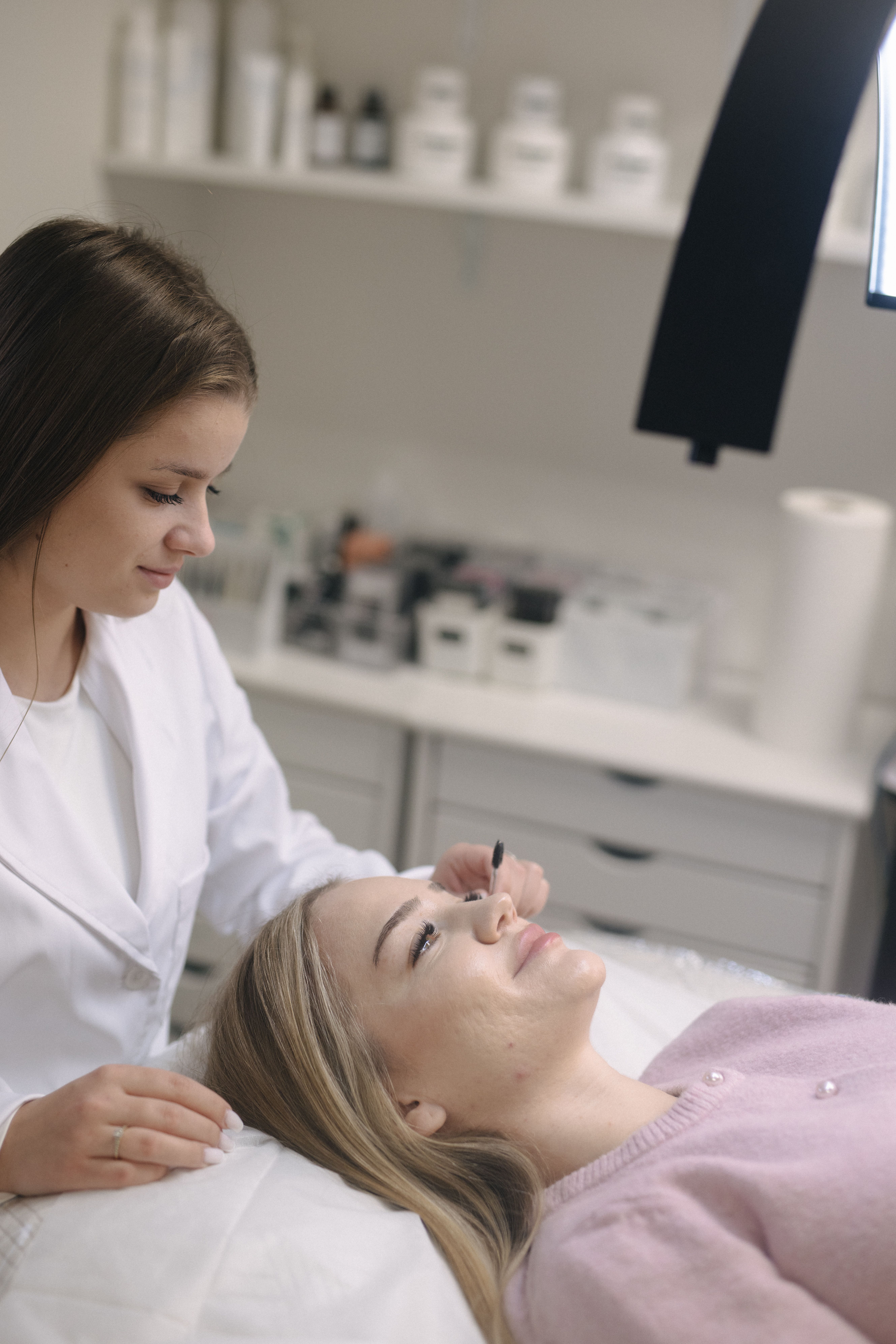 An aesthetician in a white coat grooms a client's lashes with a small brush.