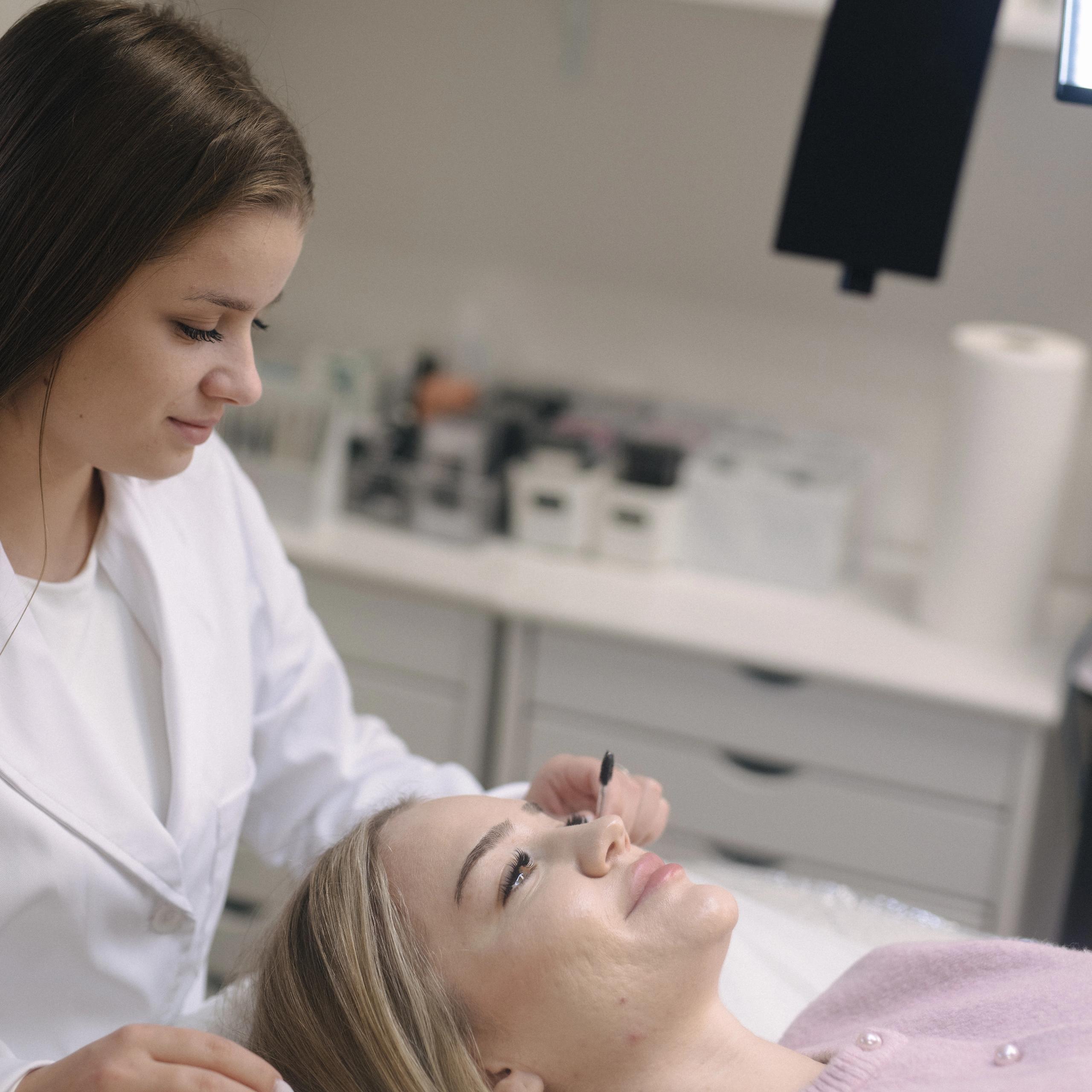 An aesthetician in a white coat grooms a client's lashes with a small brush.