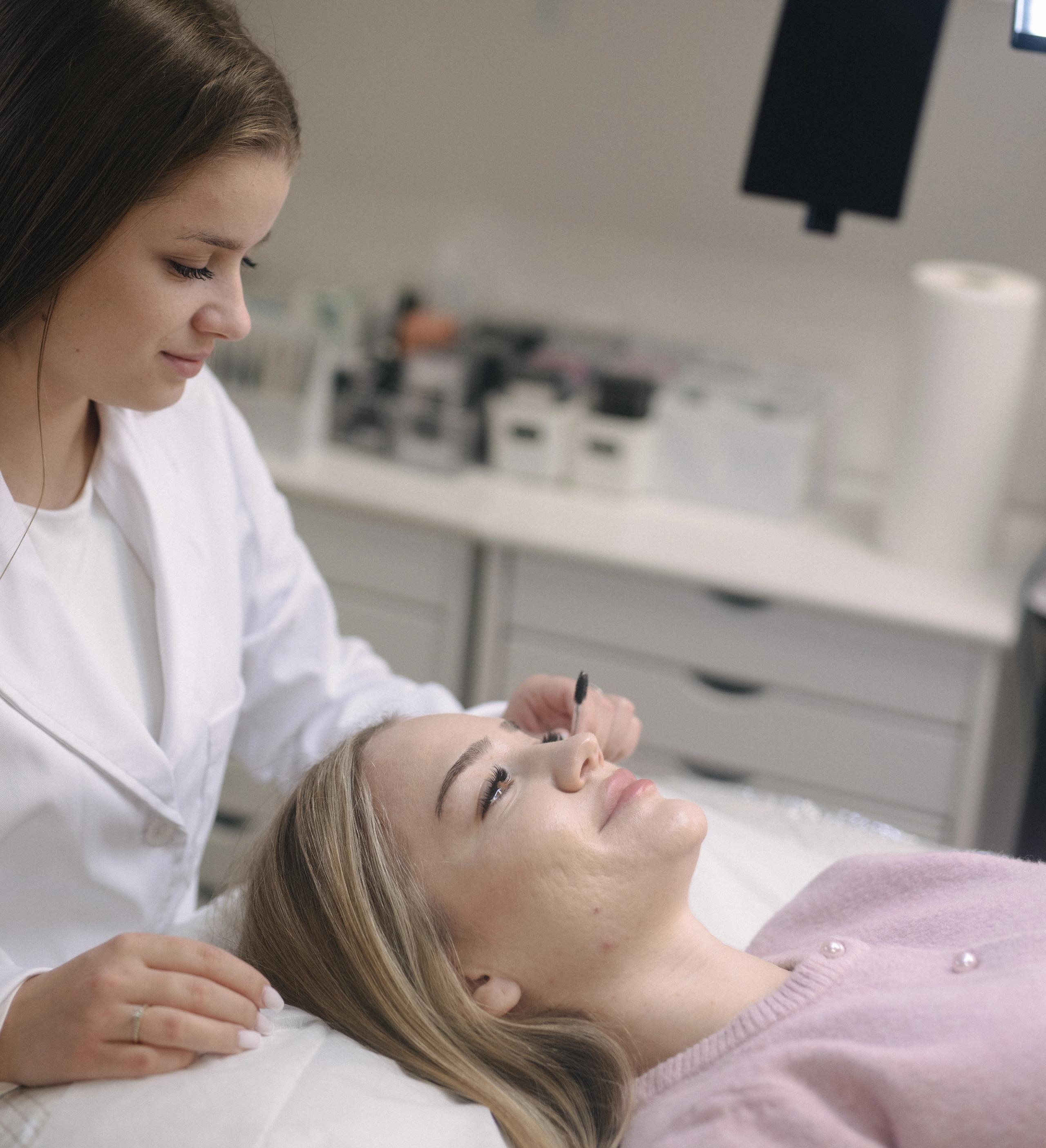 An esthetician in a white lab coat brushes a client's eyelashes.