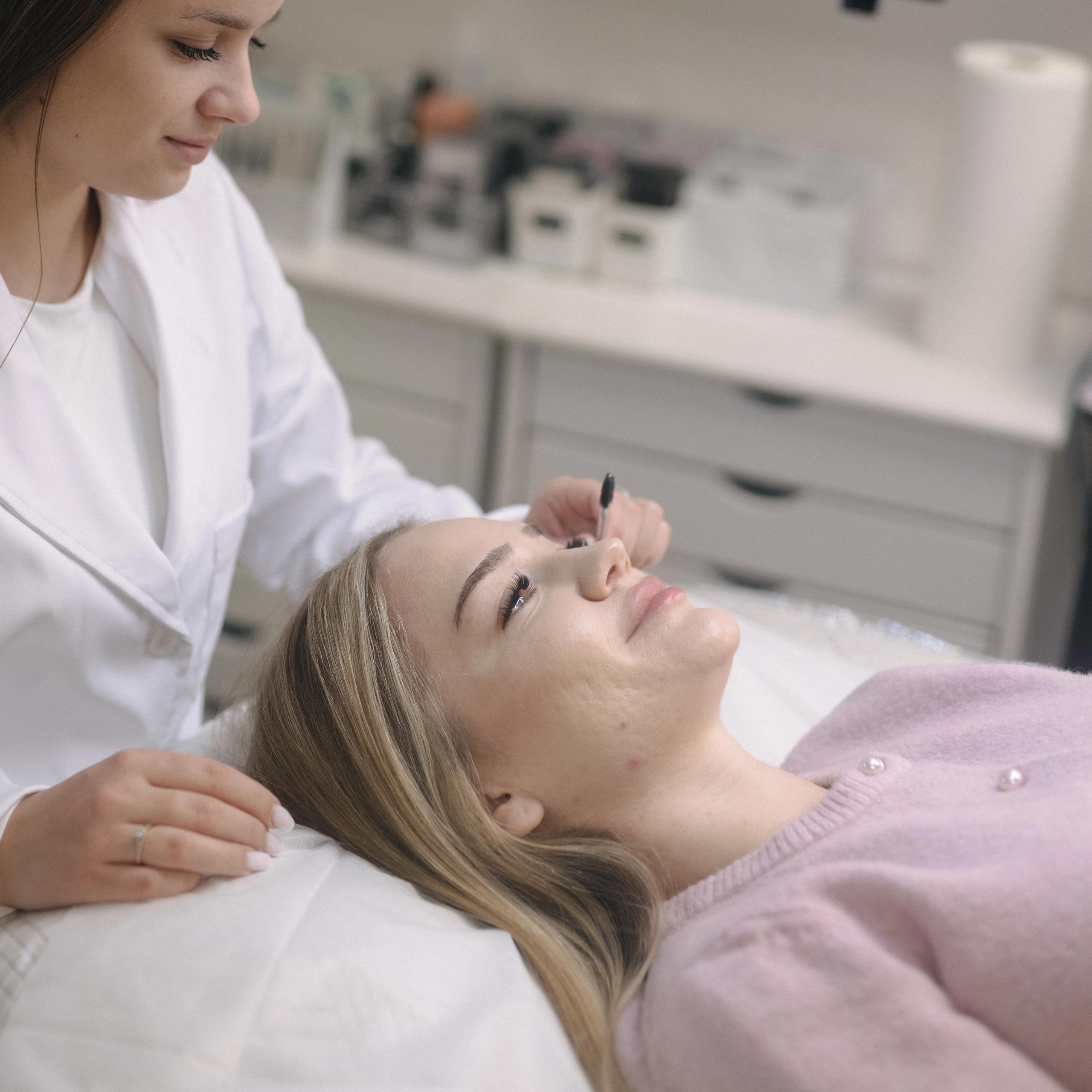 An esthetician in a white lab coat brushes a client's eyelashes.