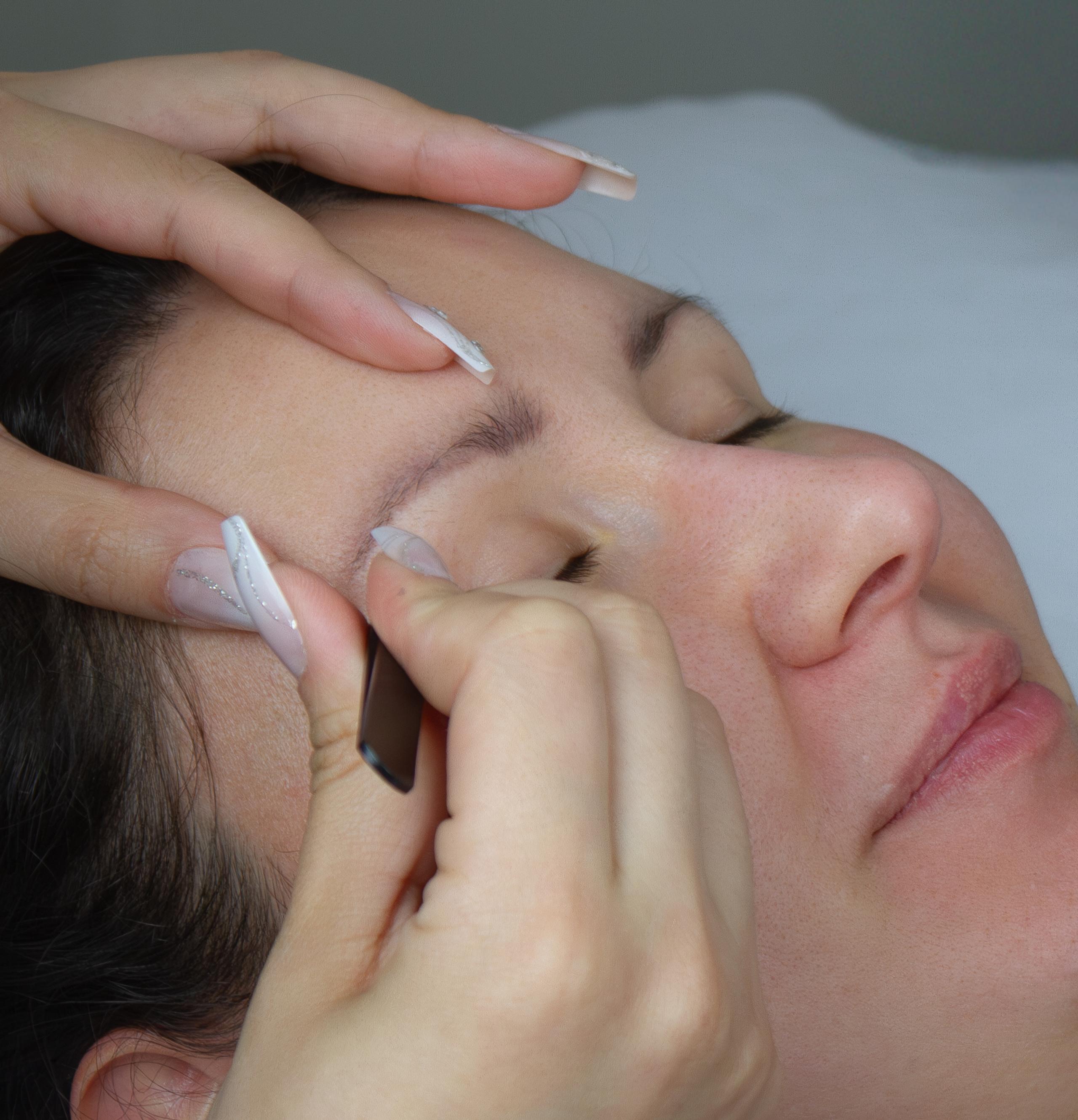 A beauty technician carefully shapes a client's eyebrow.