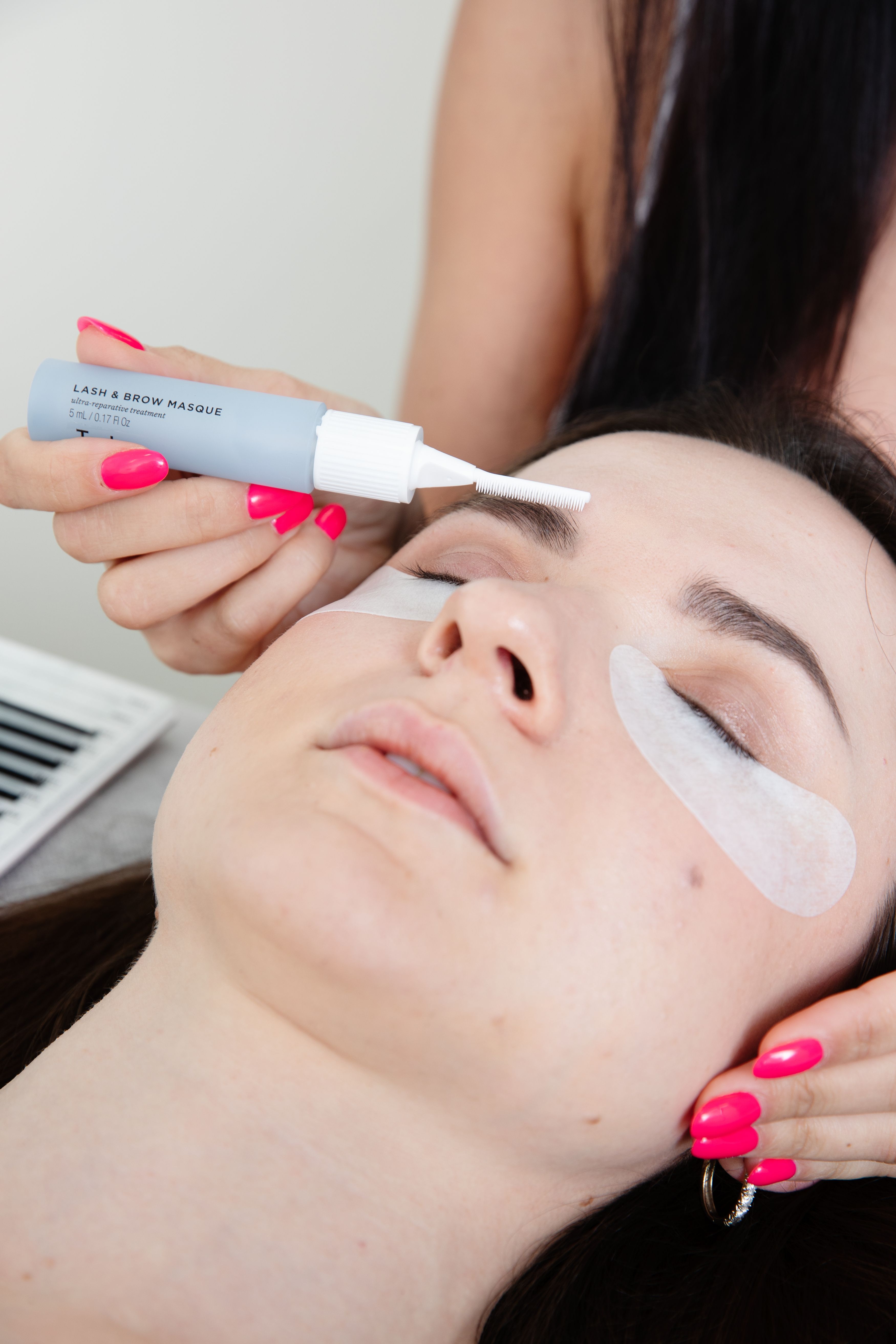 A hand with pink nails applies a "LASH & BROW MASQUE" to a person's eyebrow