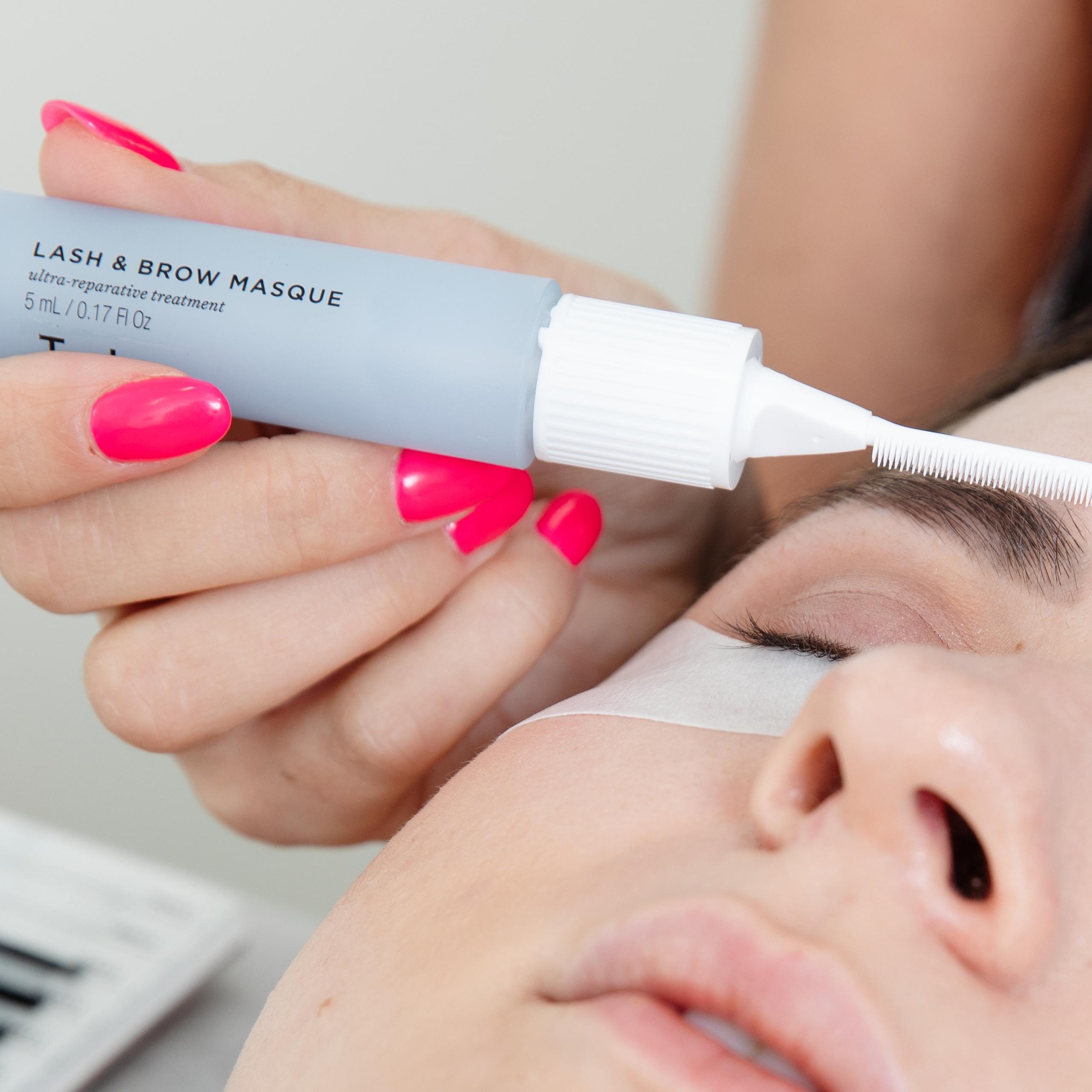 A hand with pink nails applies a "LASH & BROW MASQUE" to a person's eyebrow