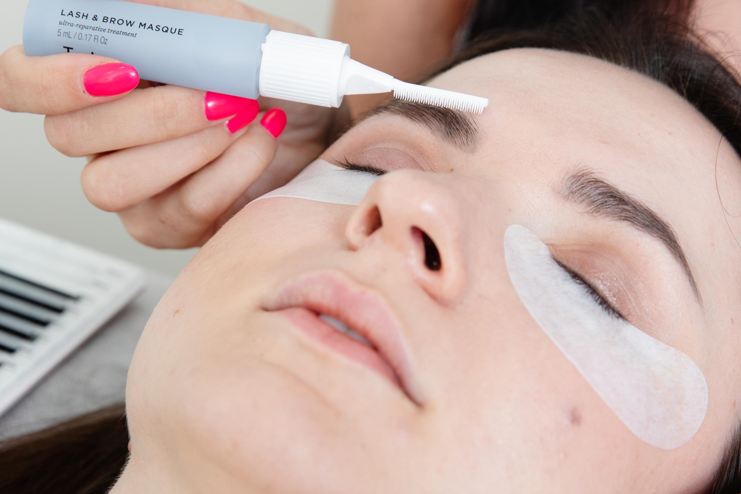 A person applies "Lash & Brow Masque" to a client's eyebrow, who has eye pads on.