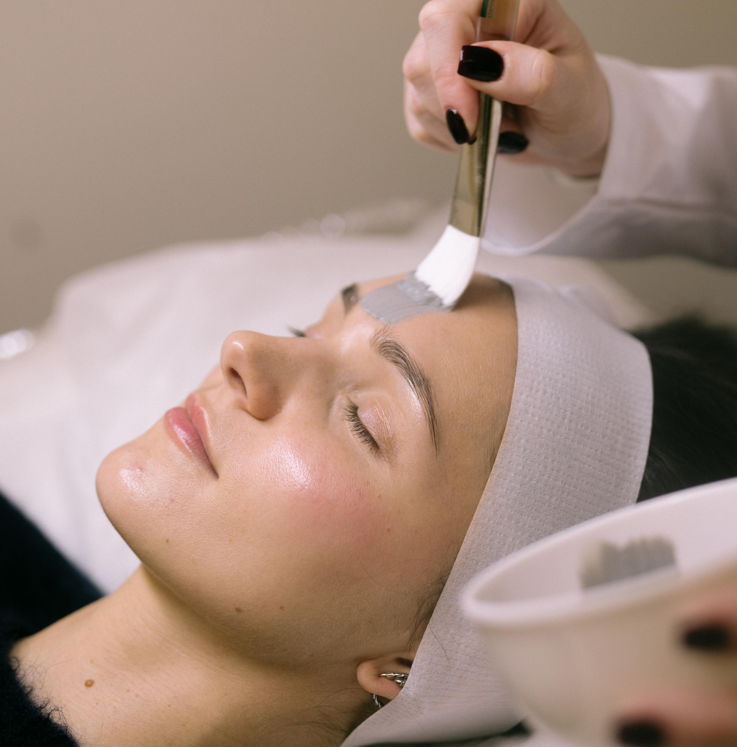 Esthetician brushing a gray mask onto a client’s forehead during a facial.
