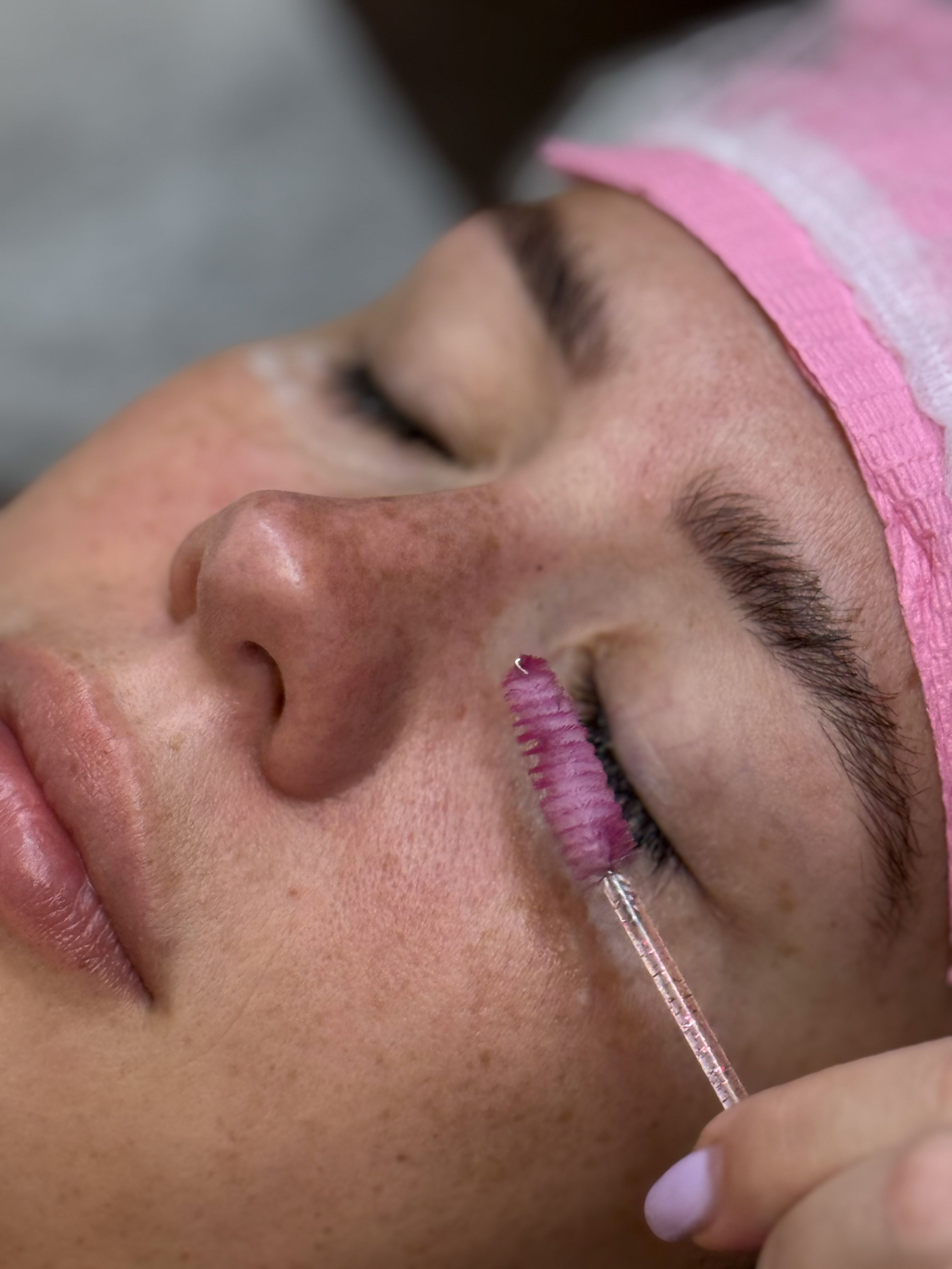 Esthetician brushing a client's lashes with a disposable lash wand while the client's eyes stay closed.