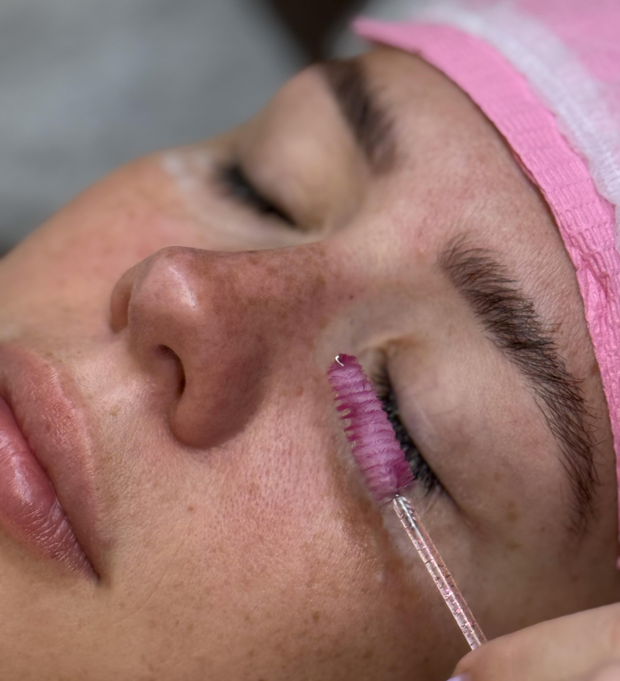 Esthetician brushing a client's lashes with a disposable lash wand while the client's eyes stay closed.