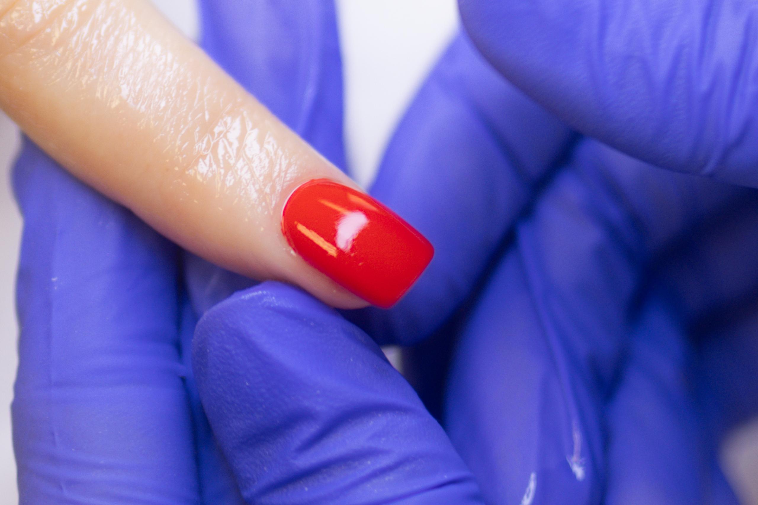 Close-up of a hand in a blue glove holding a finger with a shiny red polished nail.