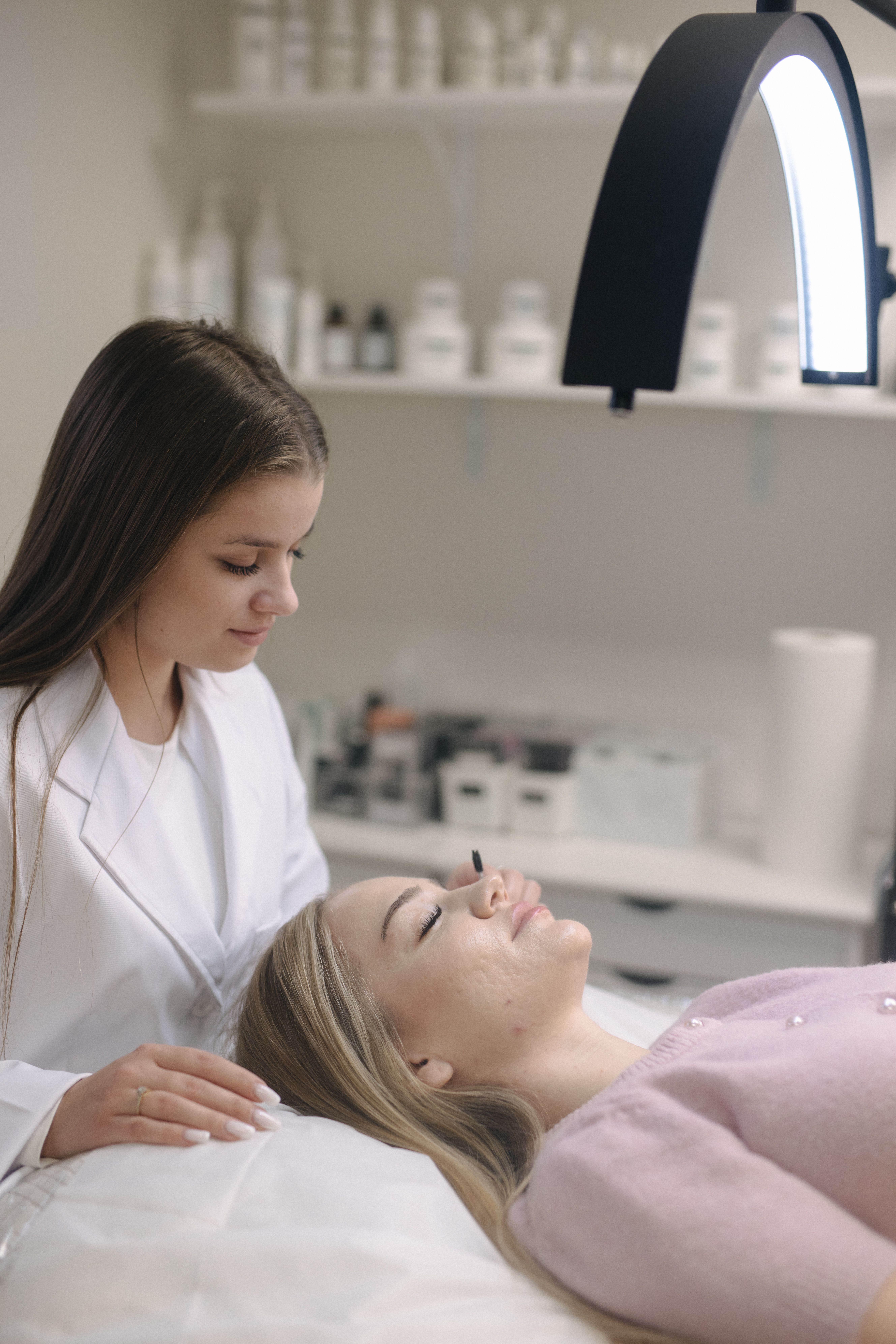 A beauty technician brushes eyelashes of a client , who lies on a bed under a bright ring light.