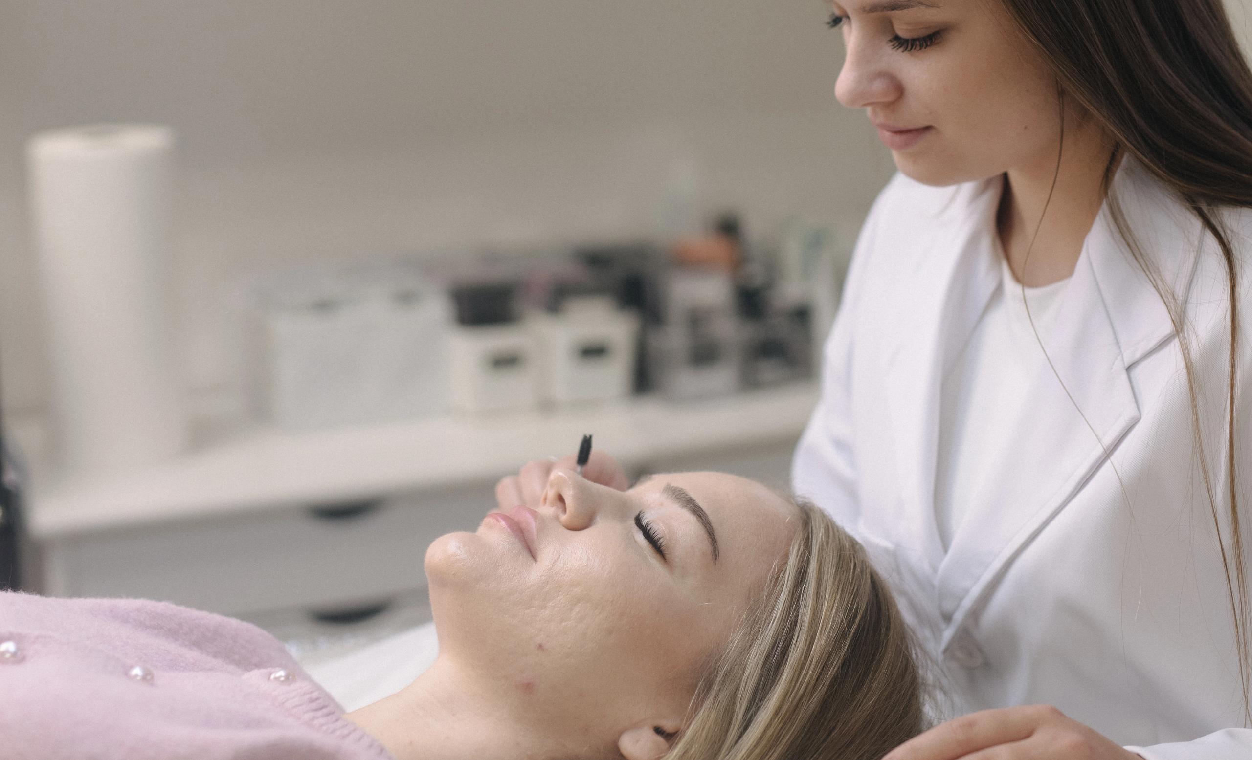 A beauty technician brushes eyelashes of a client , who lies on a bed under a bright ring light.
