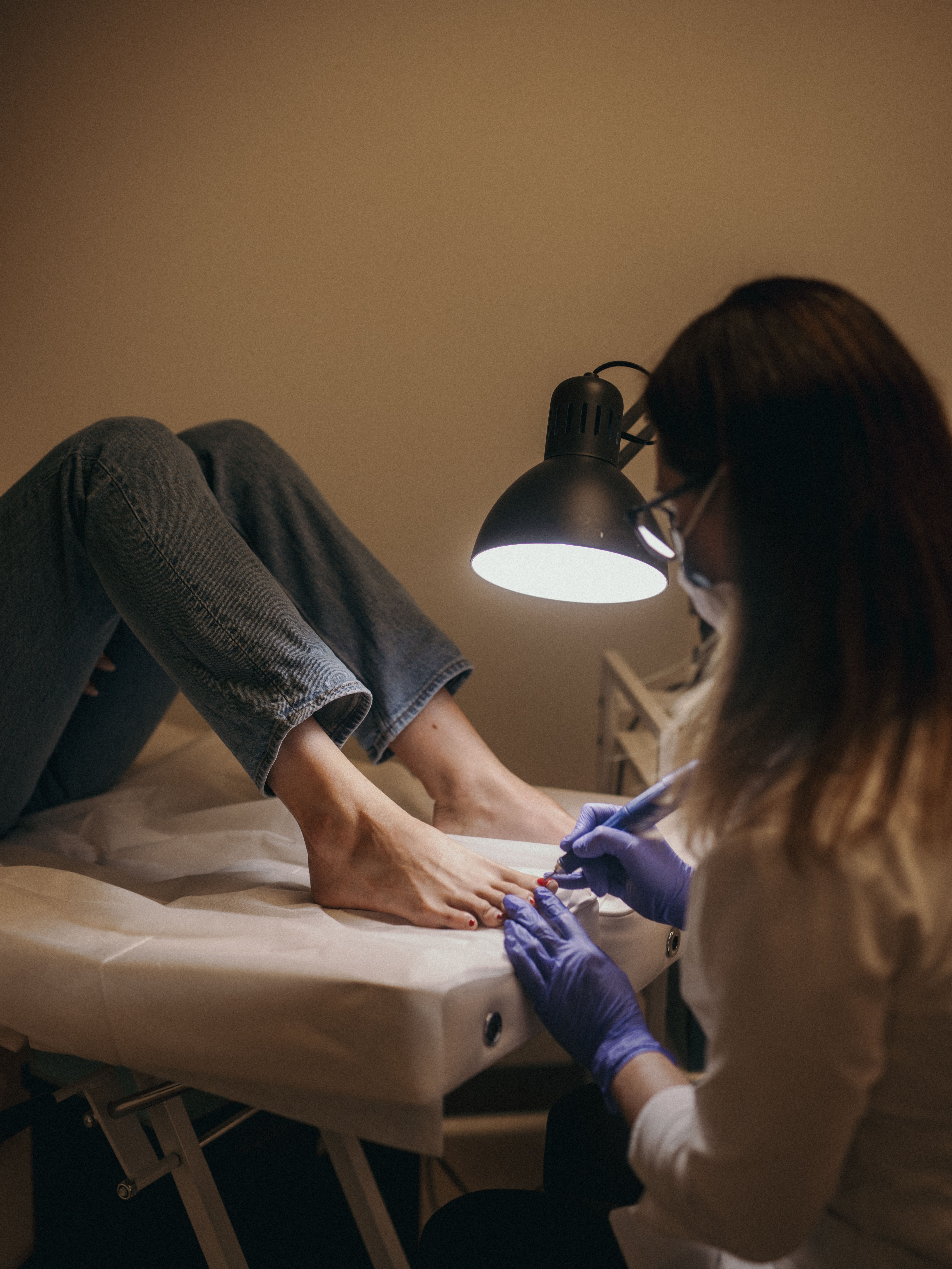 A pedicurist works on a client's feet under a bright lamp.