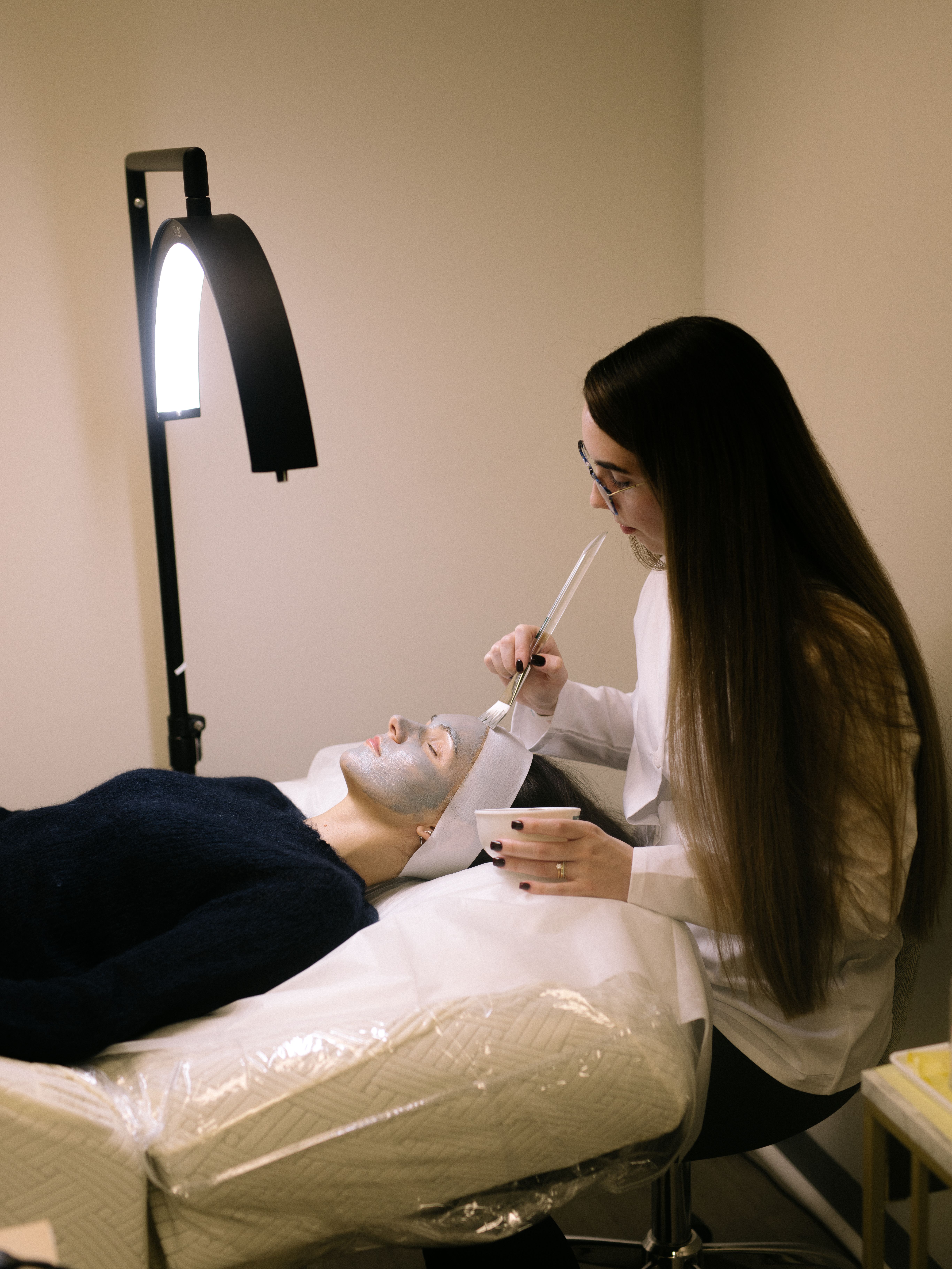 An esthetician applies a face mask to a client lying on a treatment bed.