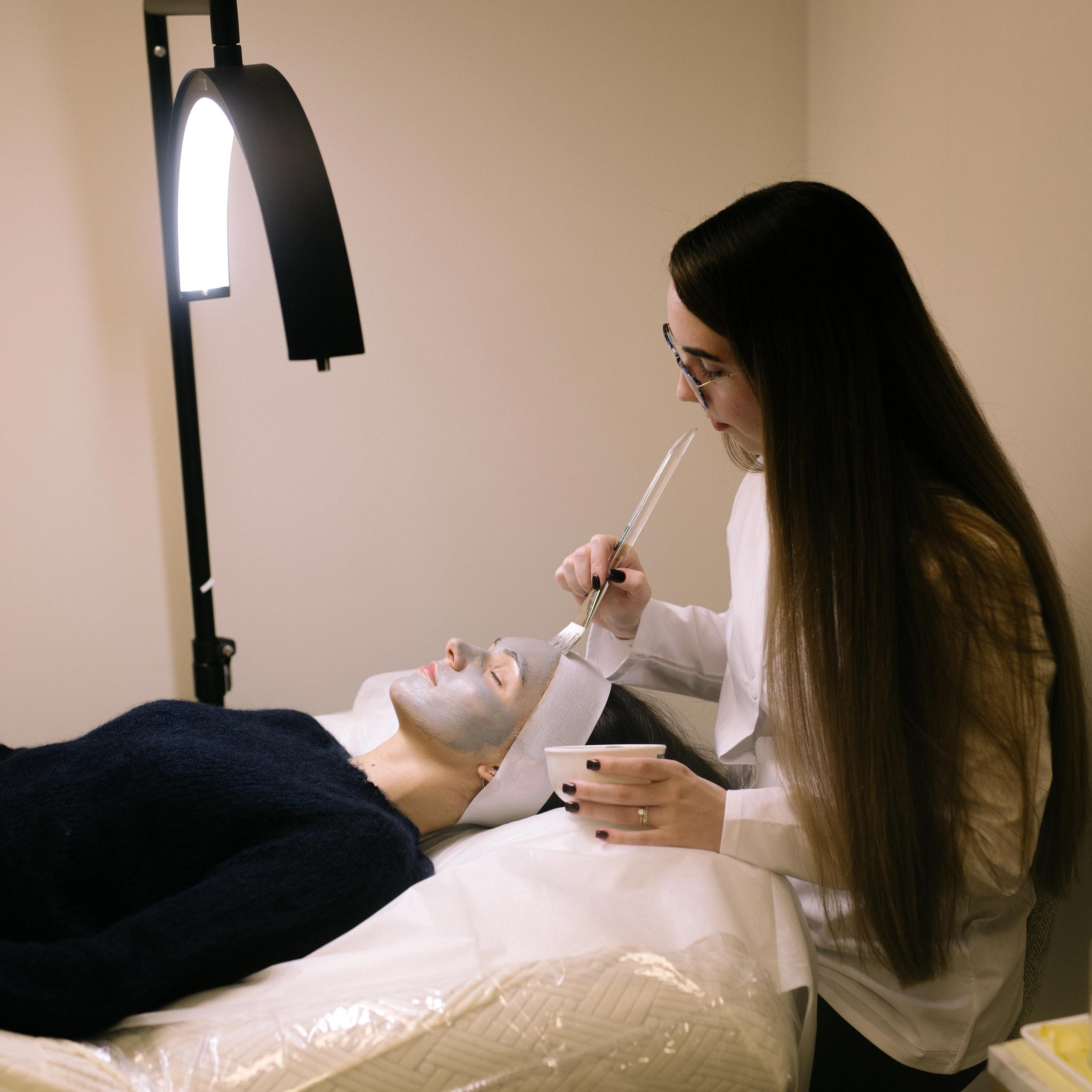 An esthetician applies a face mask to a client lying on a treatment bed.
