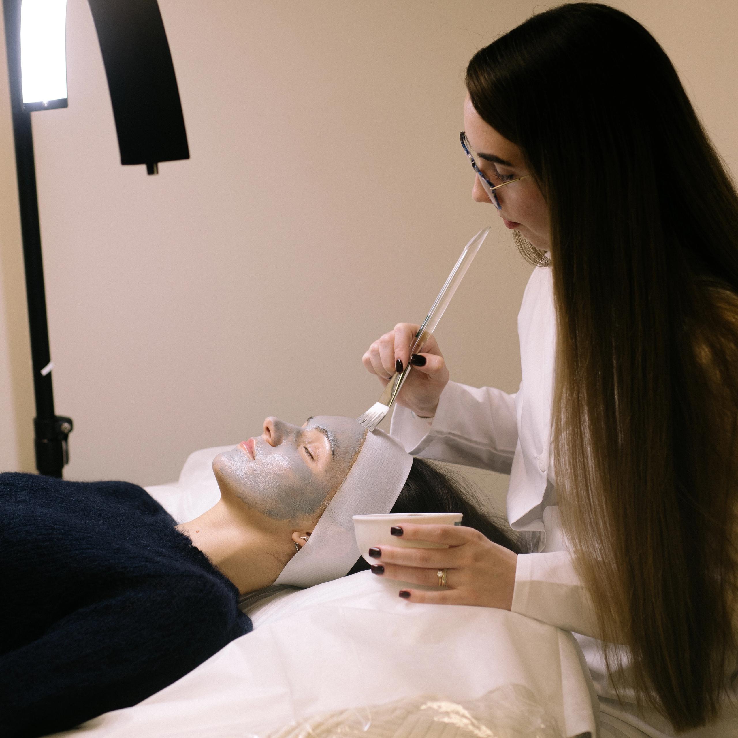 An esthetician applies a facial mask to a client lying on a treatment bed.