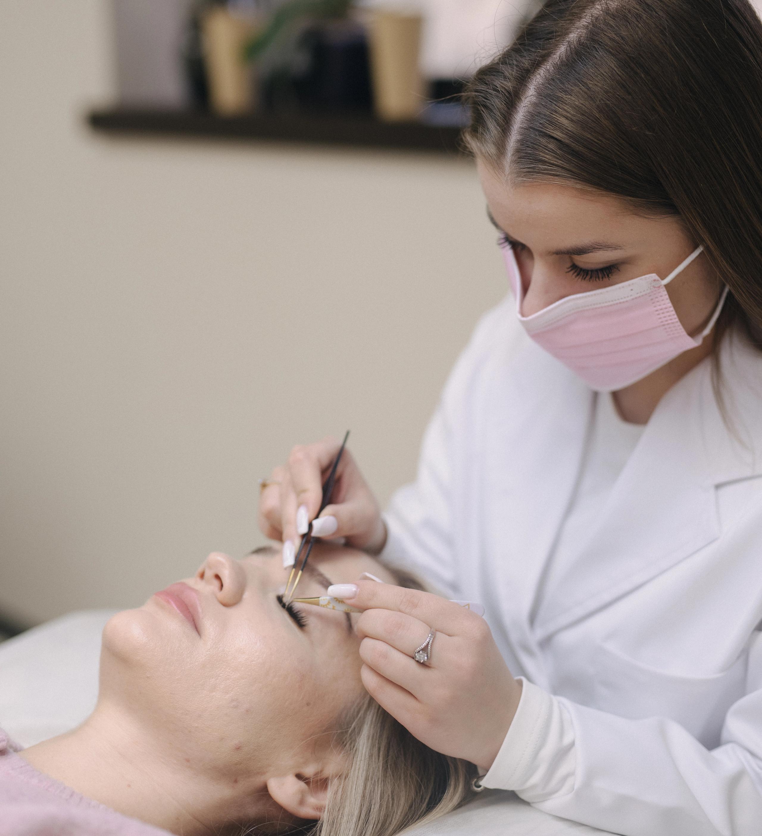 A masked technician applies eyelash extensions to a client lying on a bed.