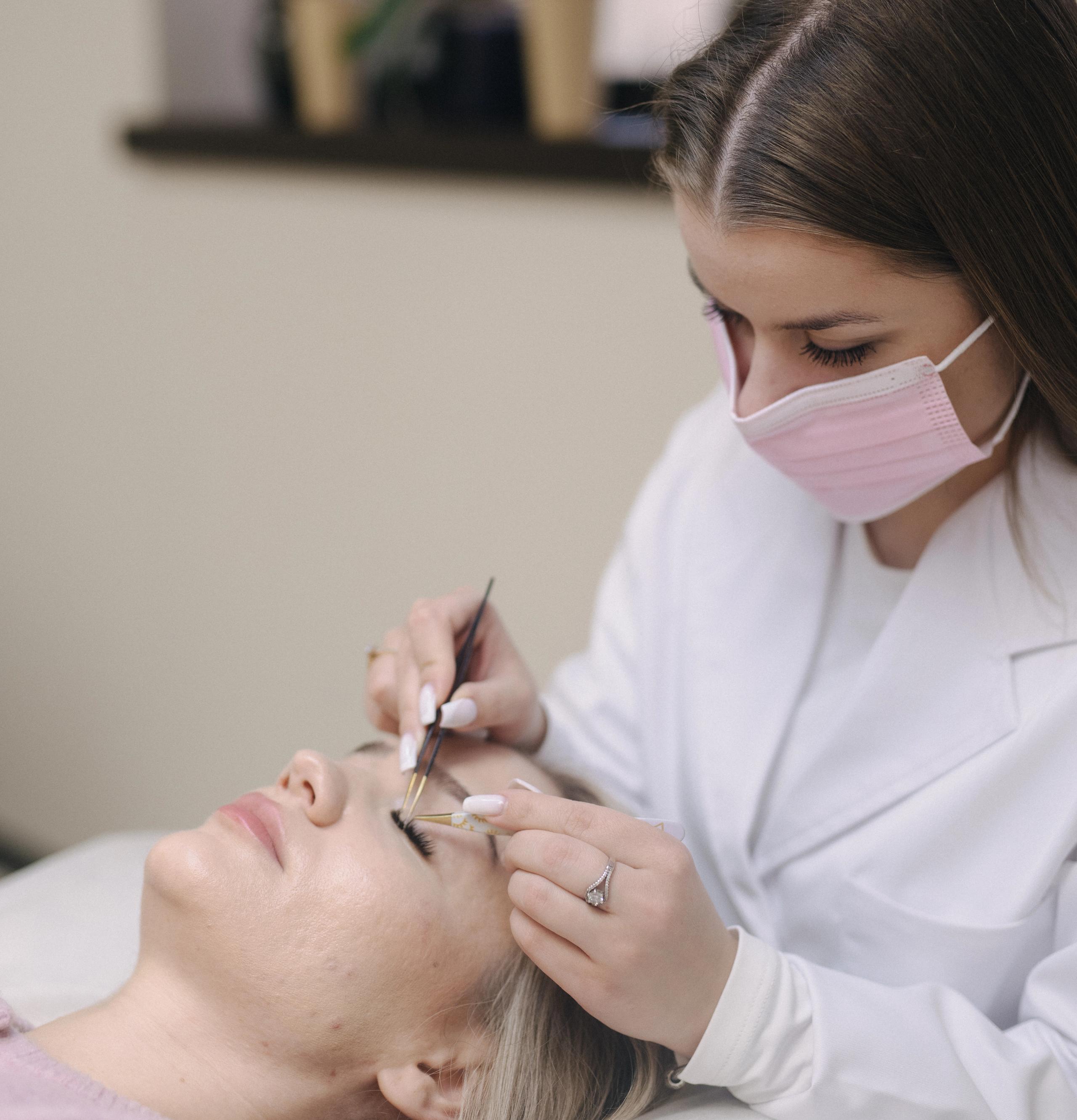 A masked technician applies eyelash extensions to a client lying on a bed.