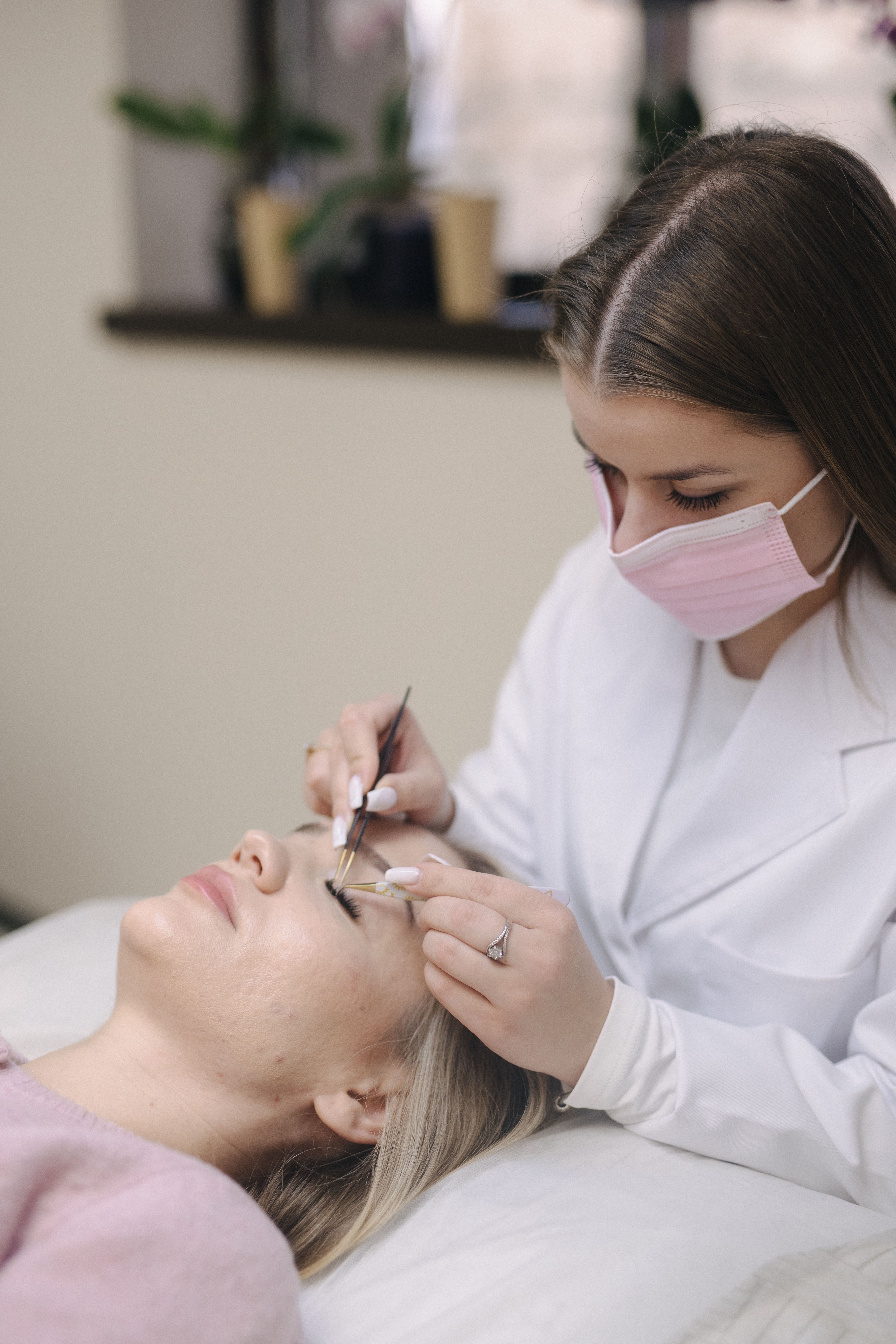 A woman receiving eyelash extensions from a masked technician.