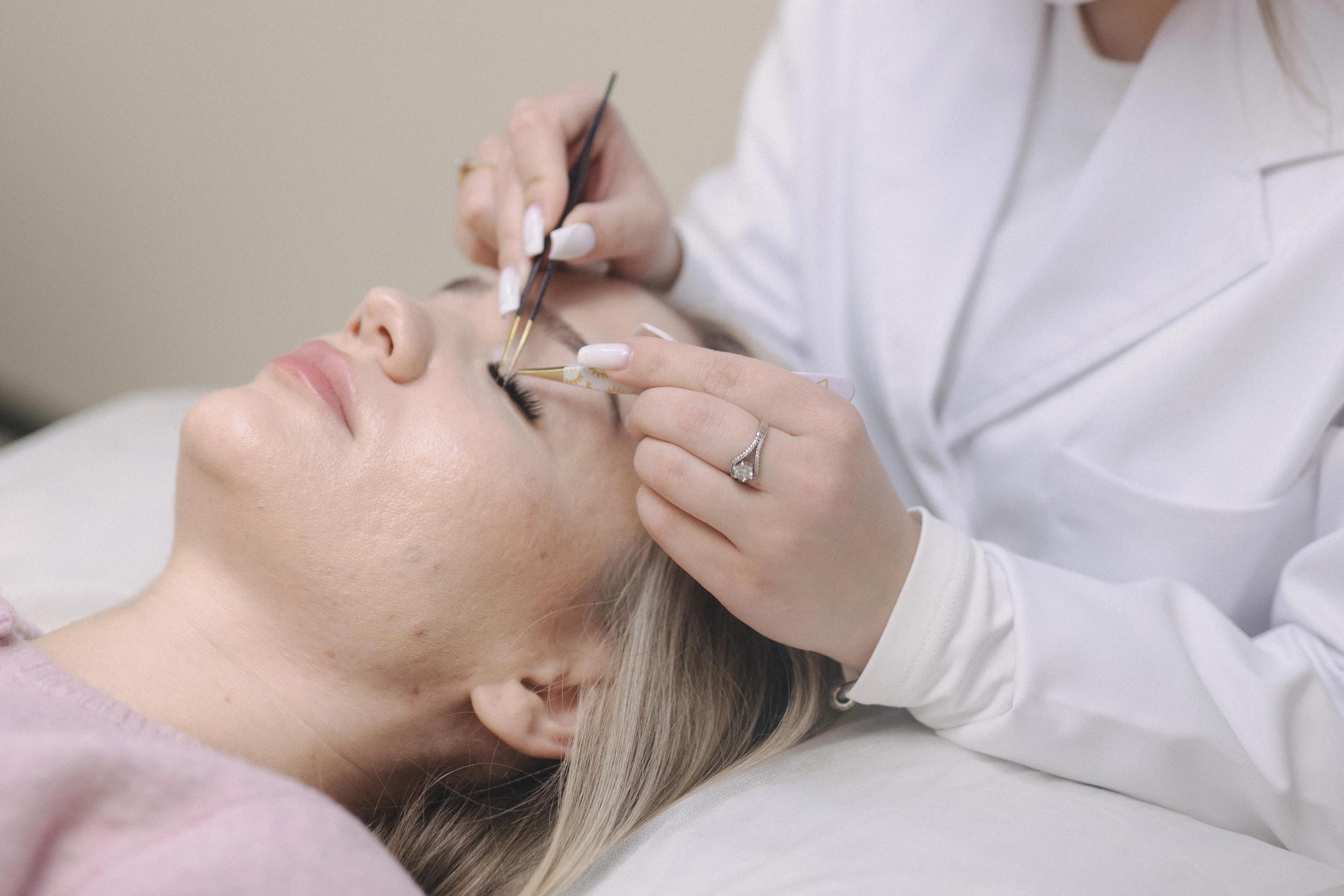 A woman receiving eyelash extensions from a masked technician.