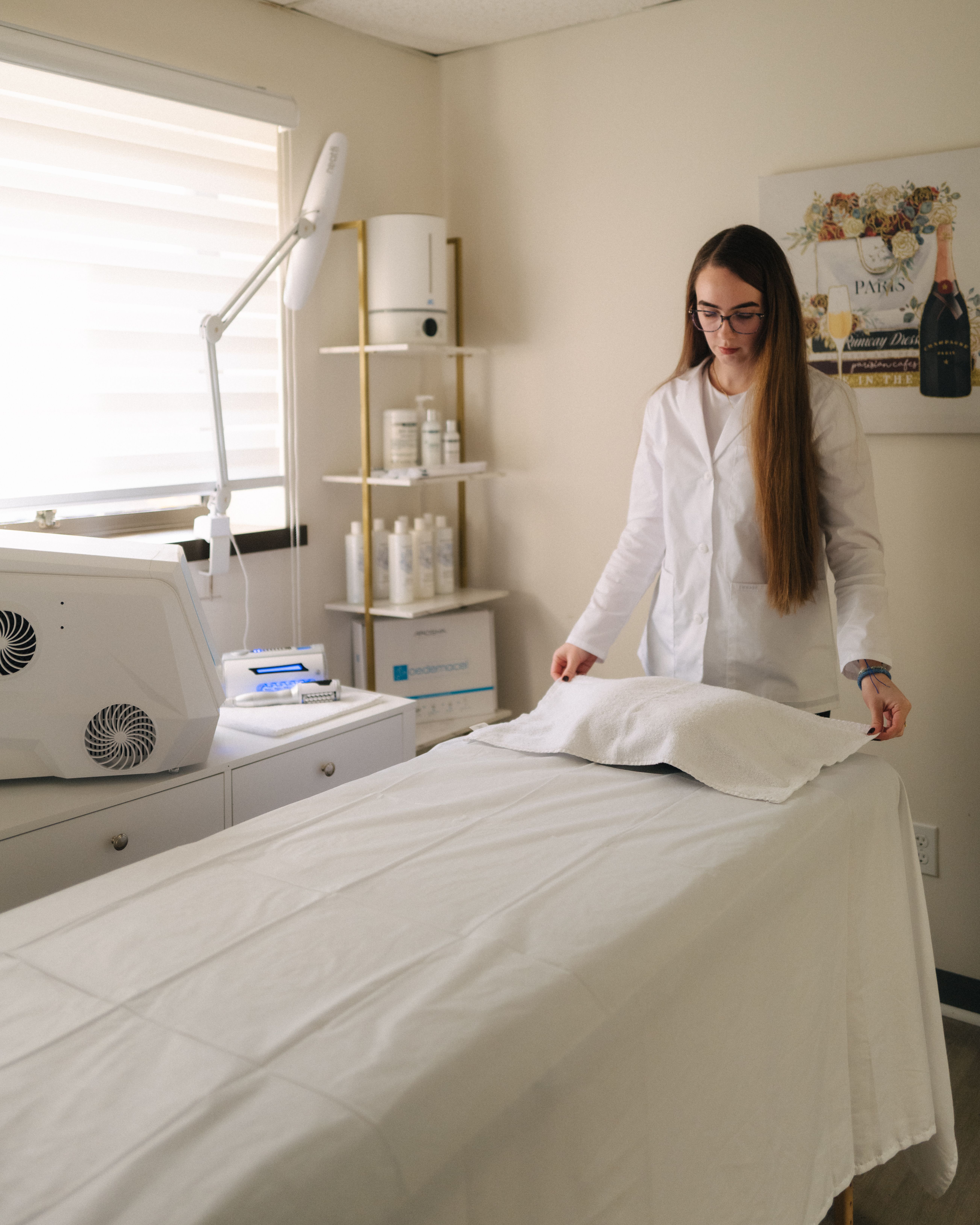 A woman in a white lab coat prepares a treatment bed in a clinic or spa room.