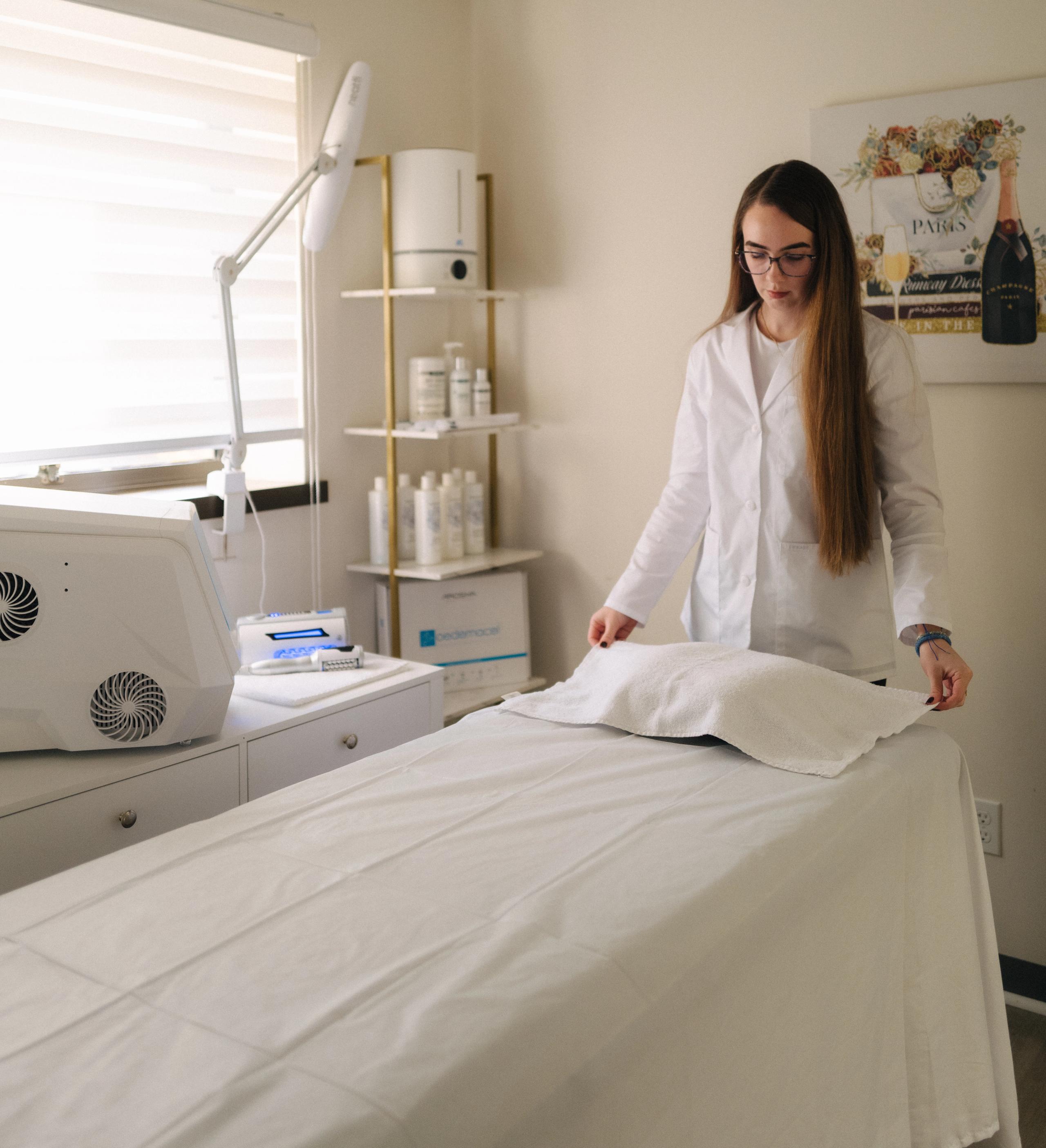 A woman in a white lab coat prepares a treatment bed in a clinic or spa room.