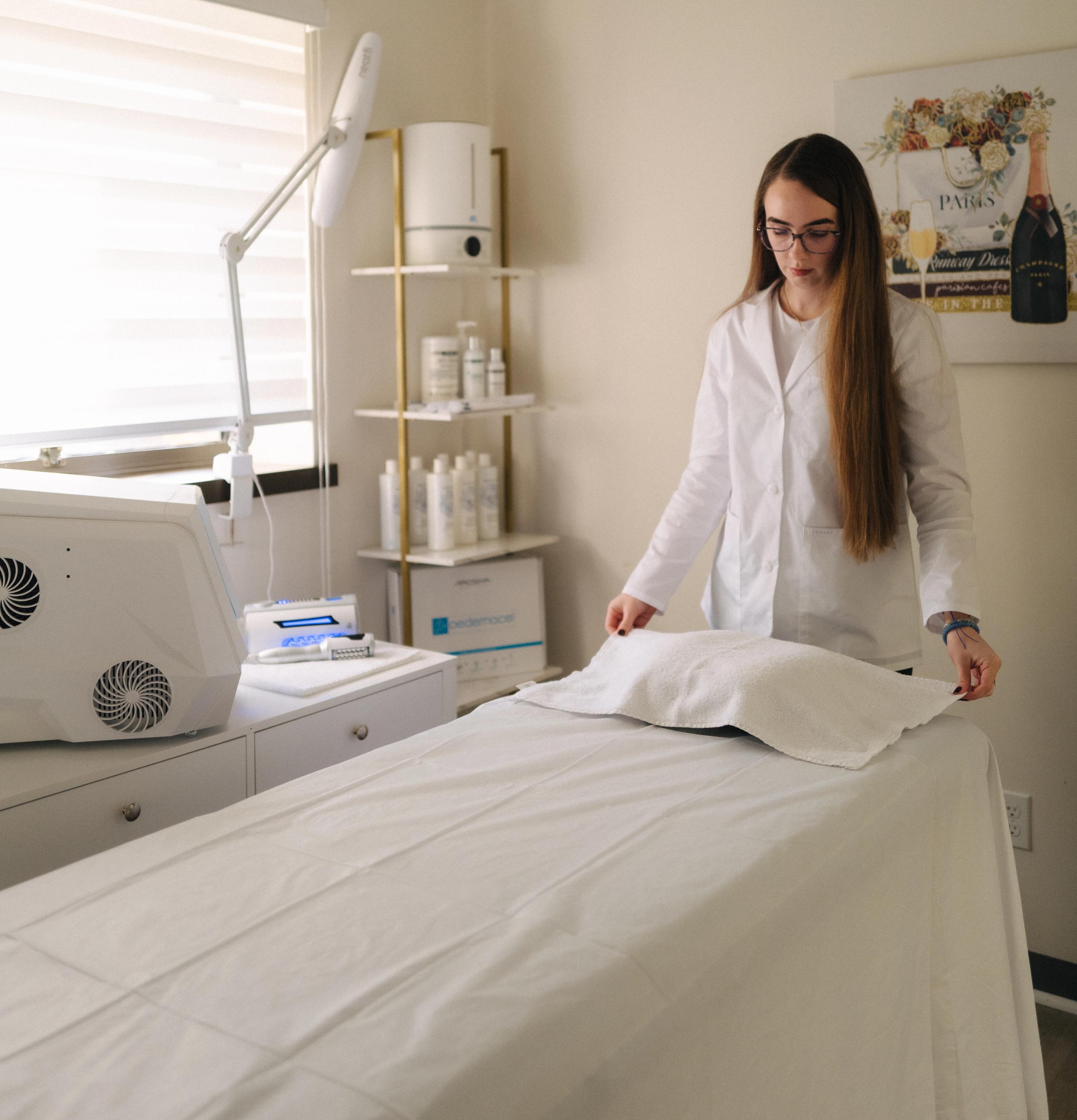 A woman in a white lab coat prepares a treatment bed in a clinic or spa room.