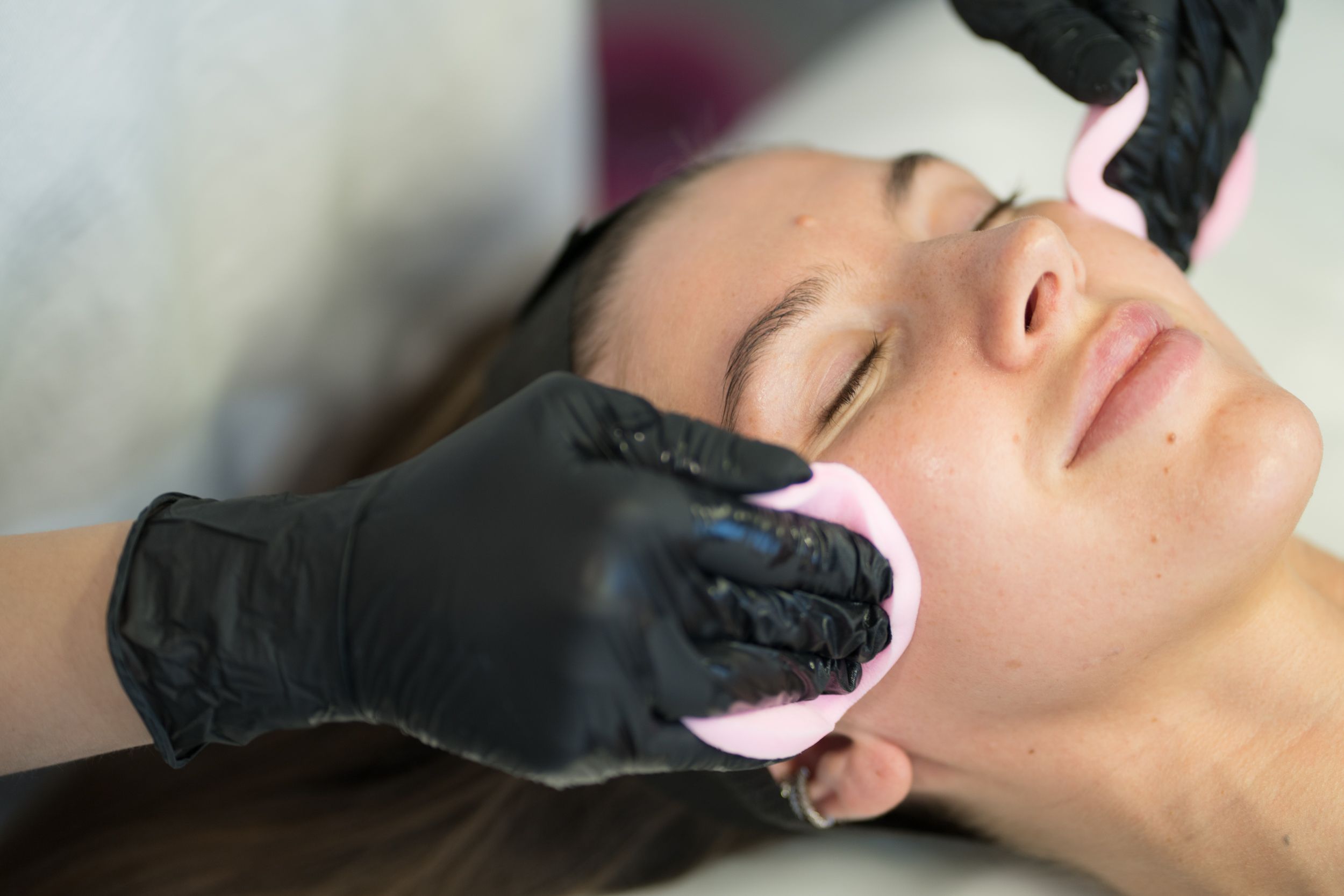Esthetician wiping a client’s face with pink sponges during a facial.