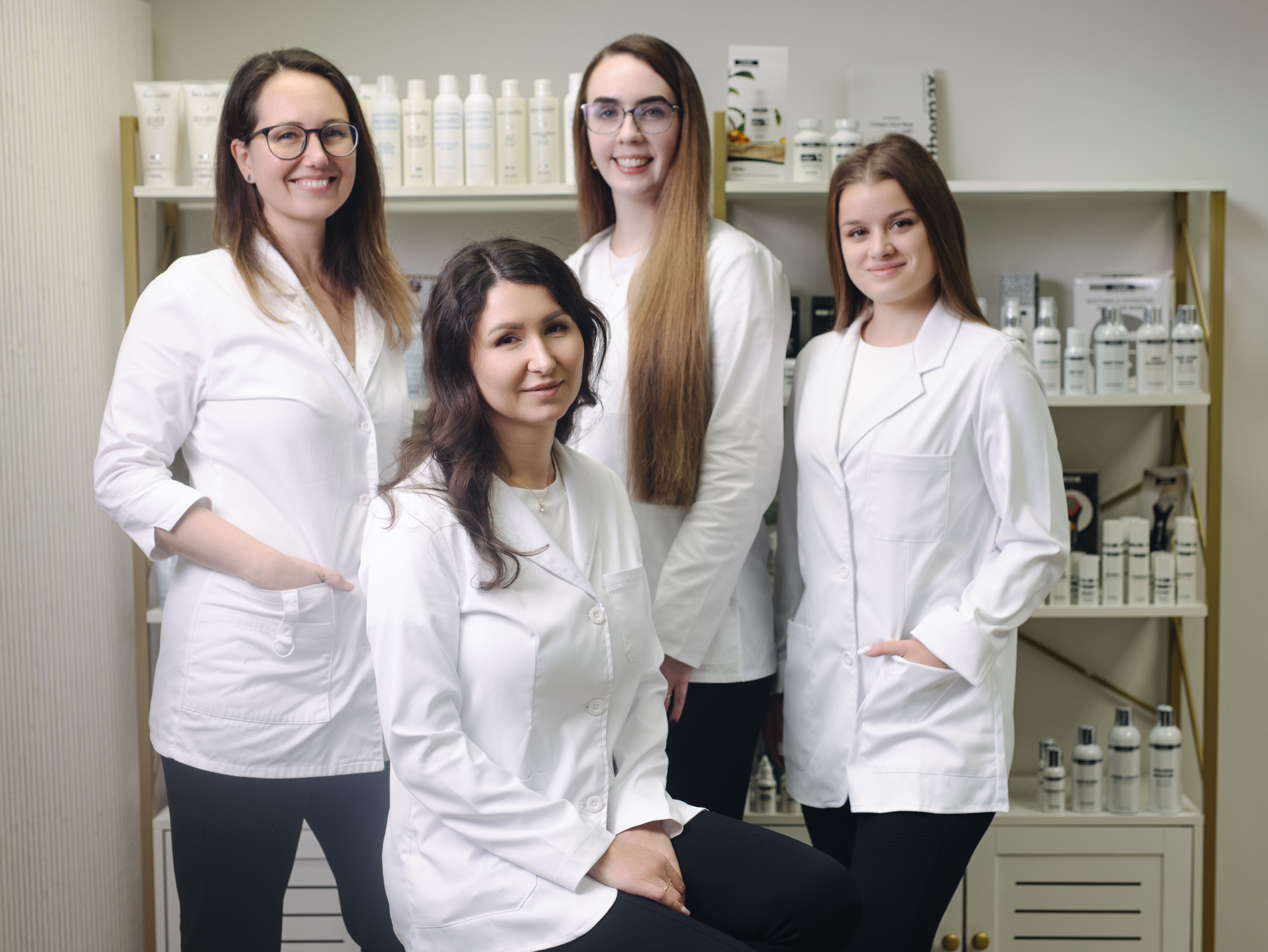 Four smiling women in white lab coats stand in front of shelves stocked with beauty products.