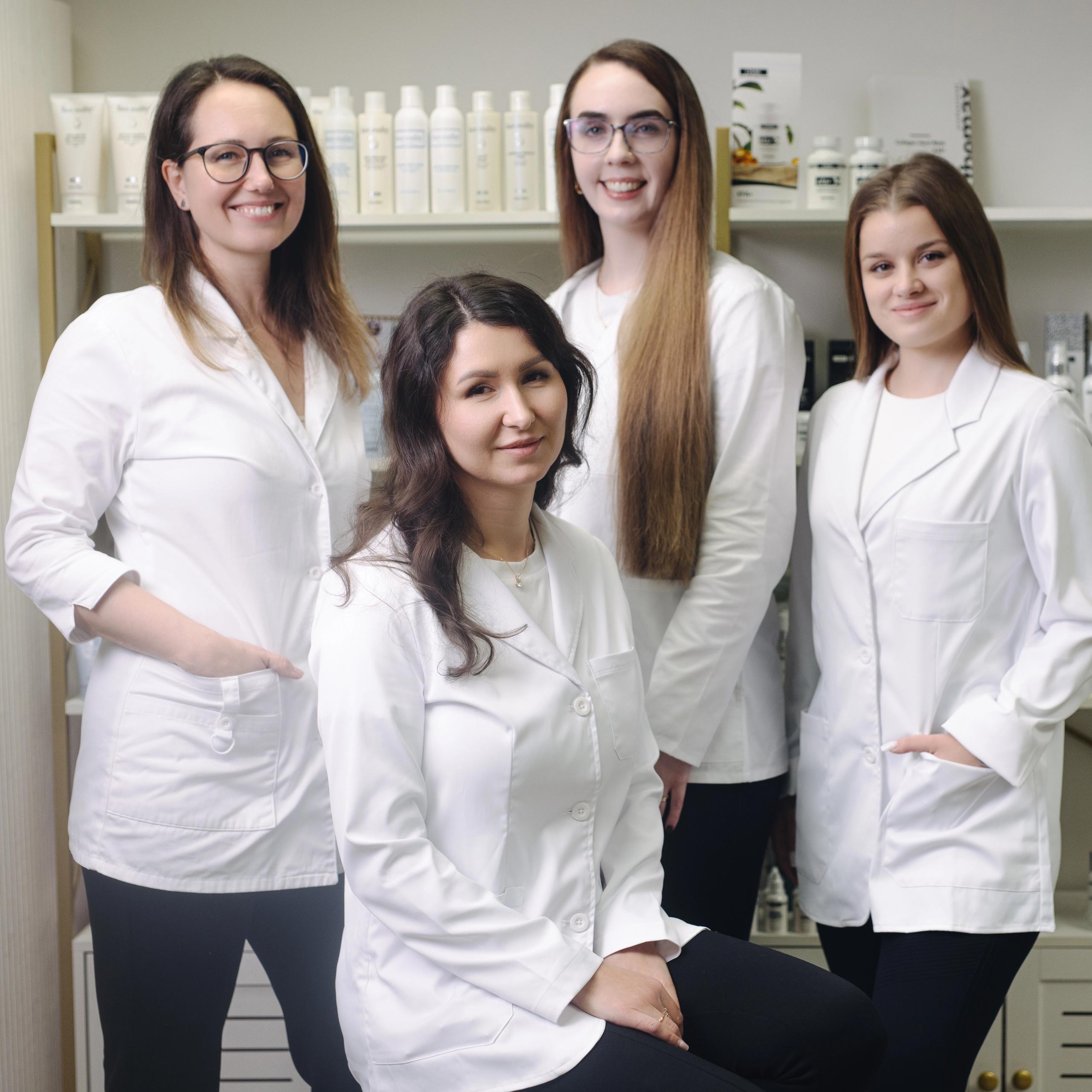 Four smiling women in white lab coats stand in front of shelves stocked with beauty products.