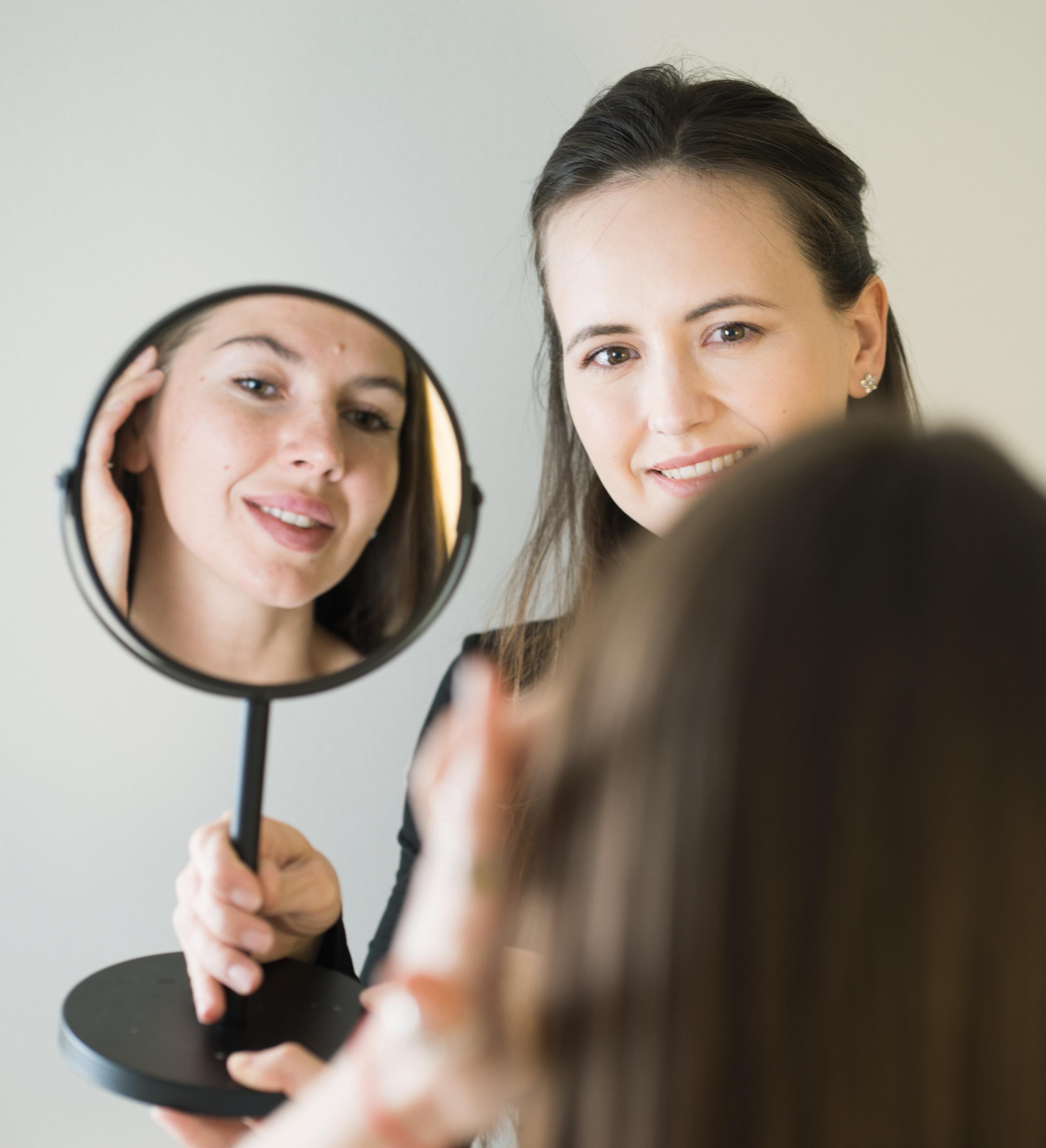 A smiling woman holds a mirror, reflecting another smiling woman touching her cheek.