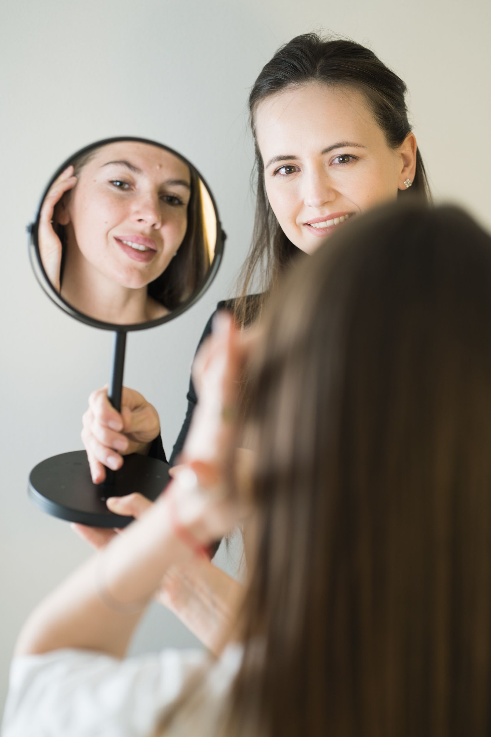 An esthetician holding a mirror, showing a client results of her brow treatment. Both are smiling 