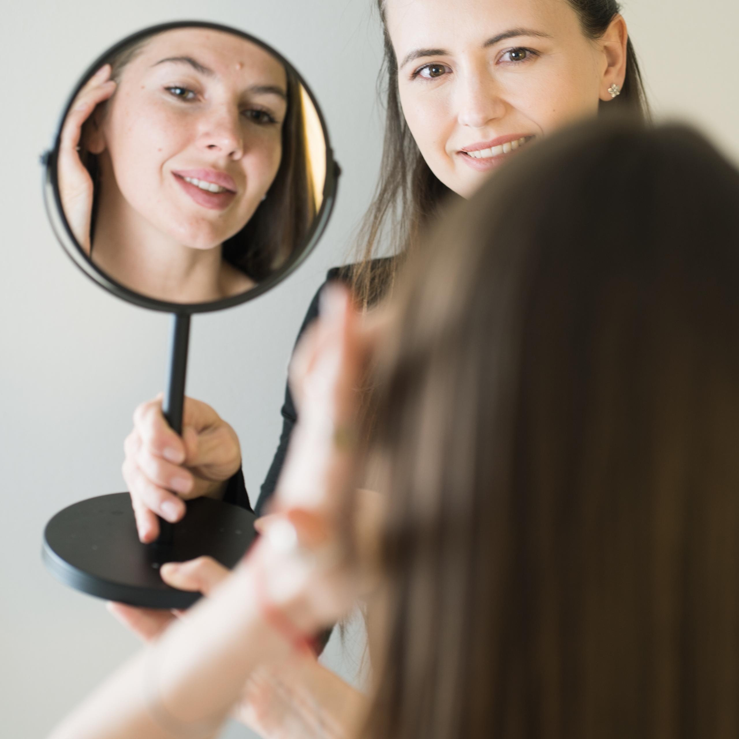 An esthetician holding a mirror, showing a client results of her brow treatment. Both are smiling