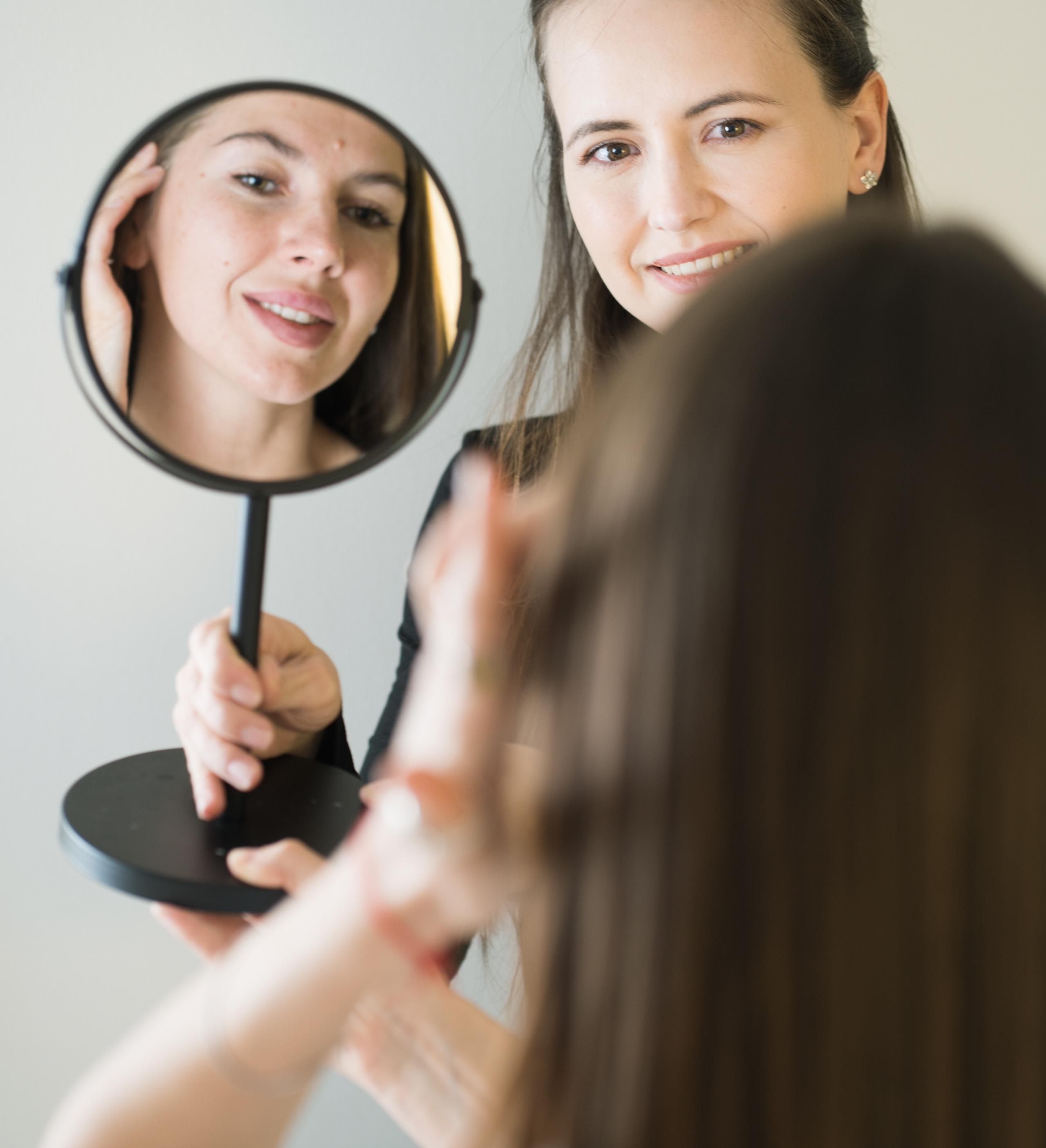 Brow tint client checks her finished brows in a hand mirror while her esthetician looks on.