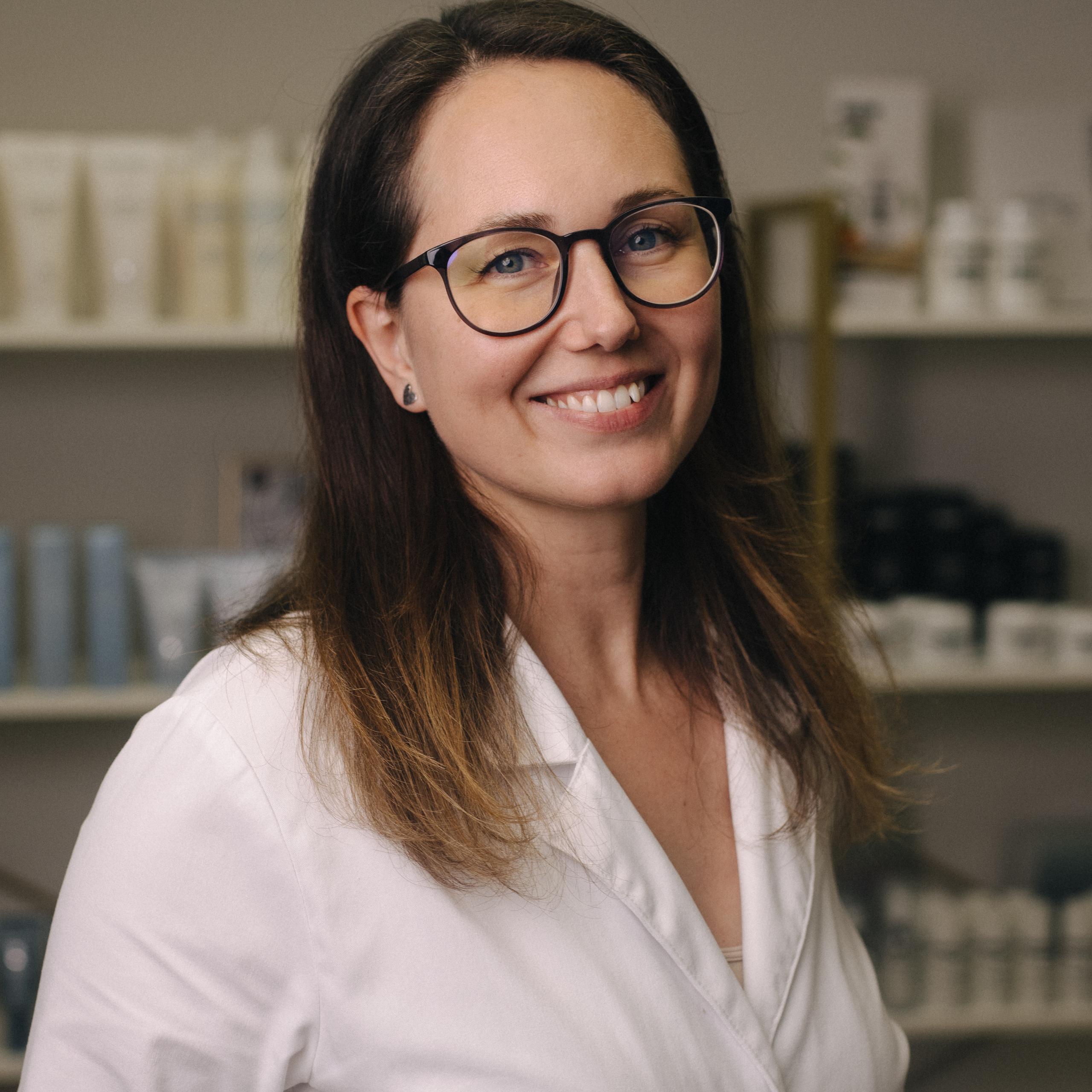 A smiling woman in a white lab coat and glasses stands in a salon waiting room