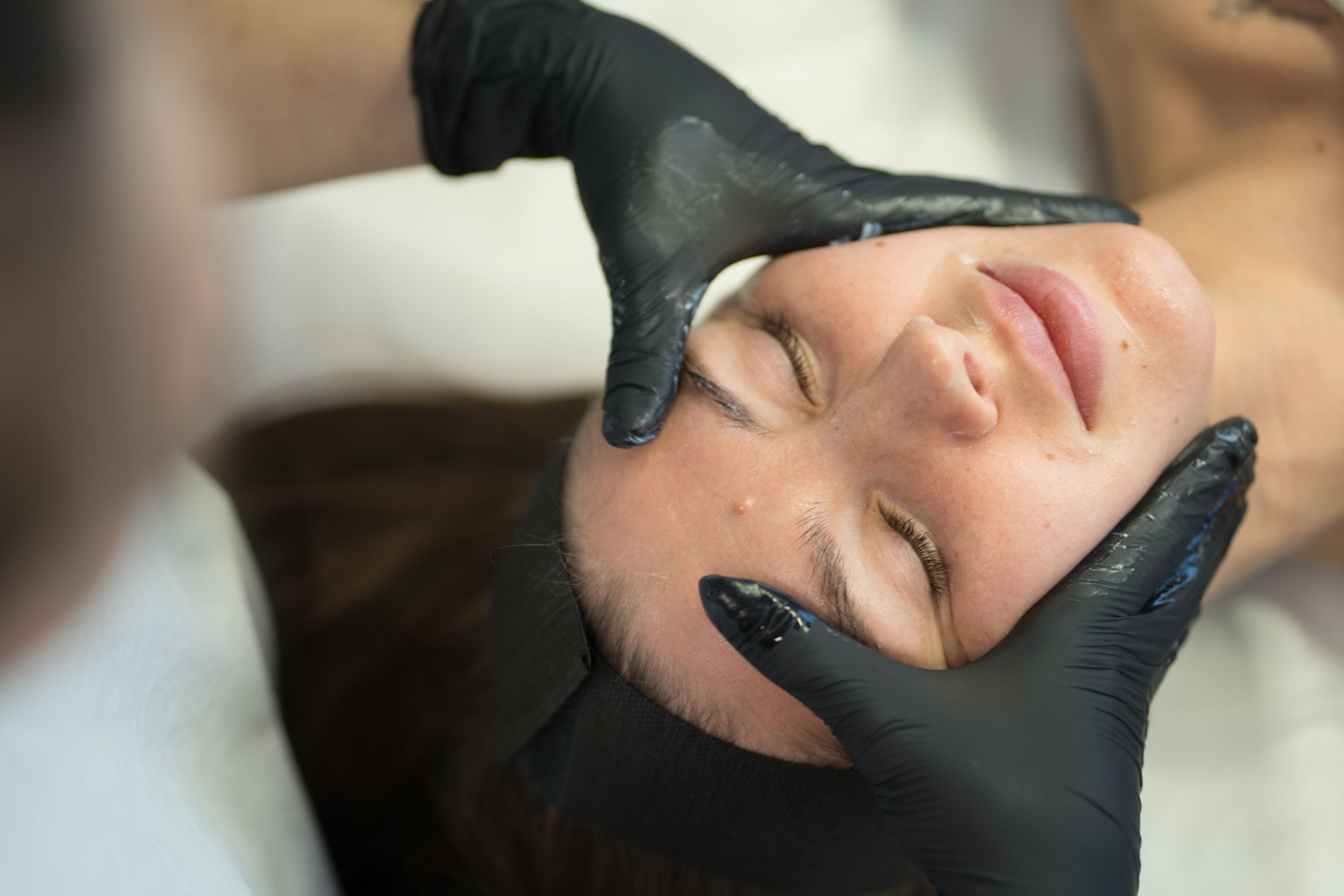 A person with closed eyes receives a facial treatment from gloved hands.