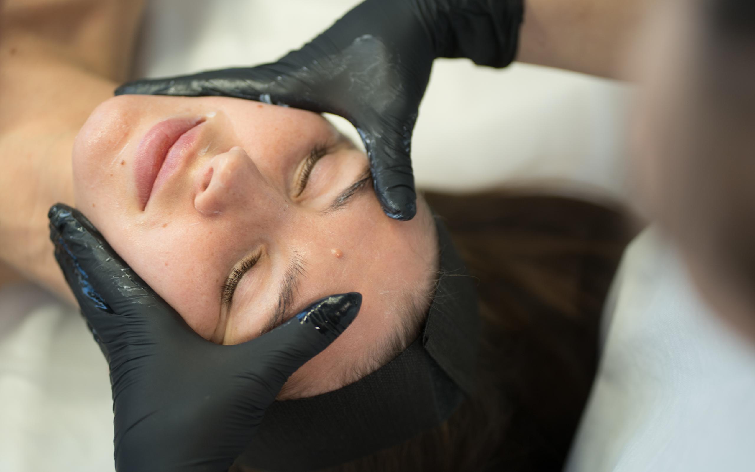 A person with closed eyes receives a facial treatment from gloved hands.