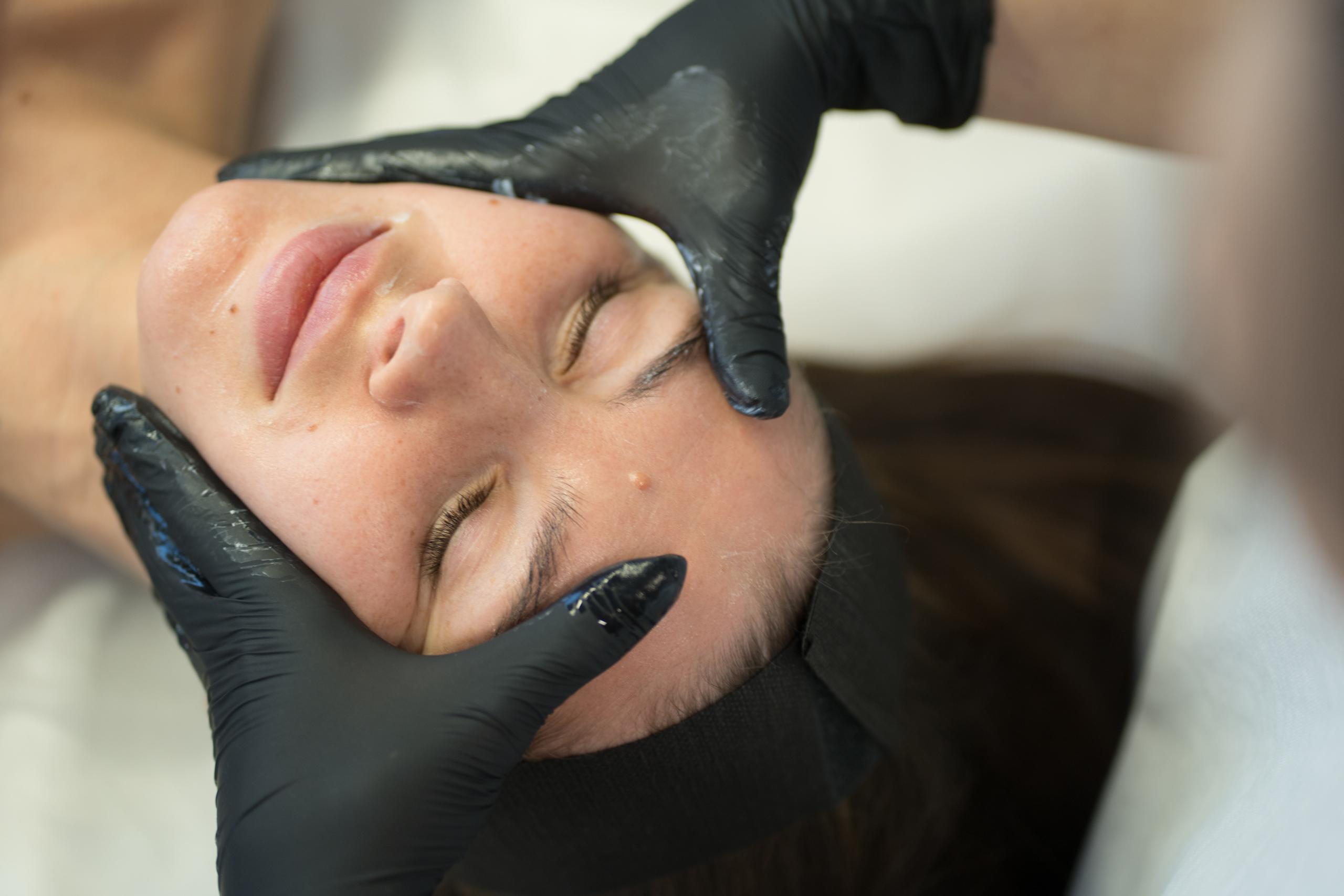 A person with closed eyes receives a facial treatment from gloved hands.