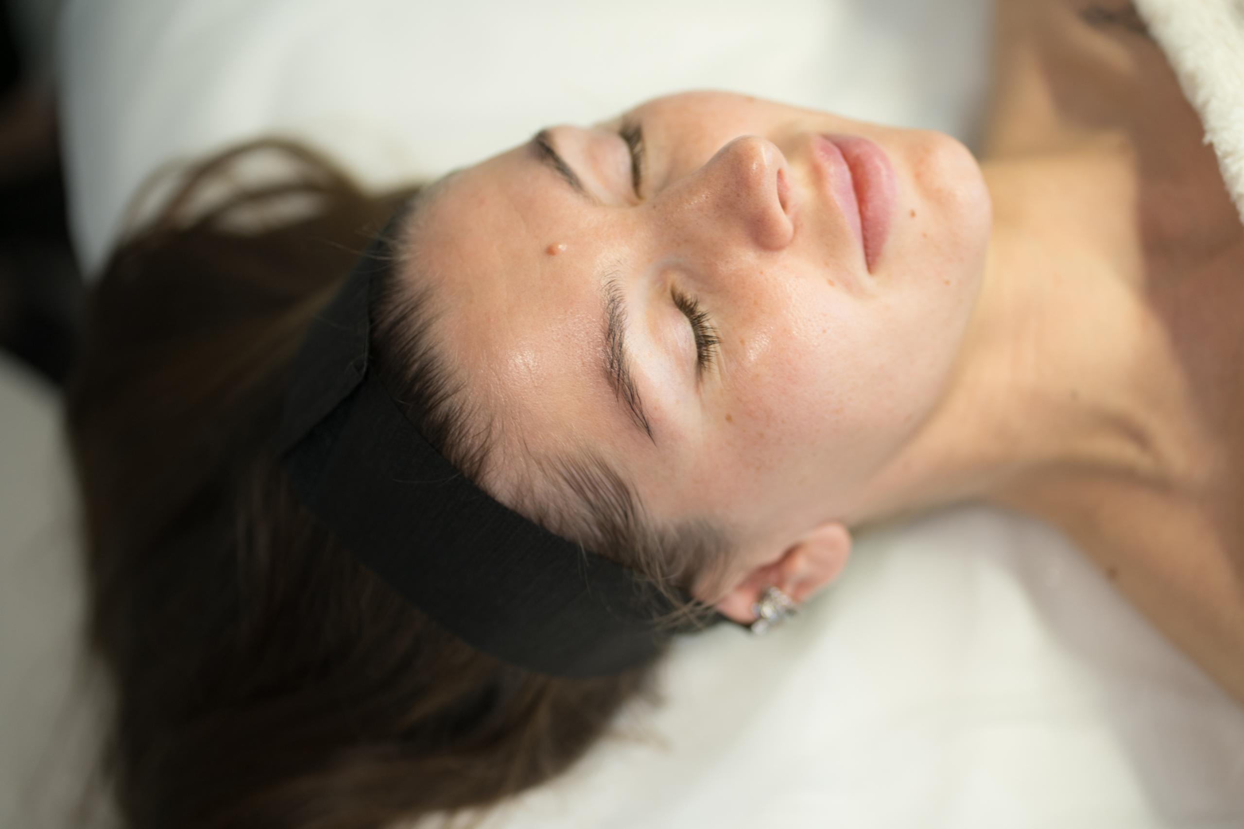 A woman with closed eyes wearing a black headband, relaxing on a white bed.