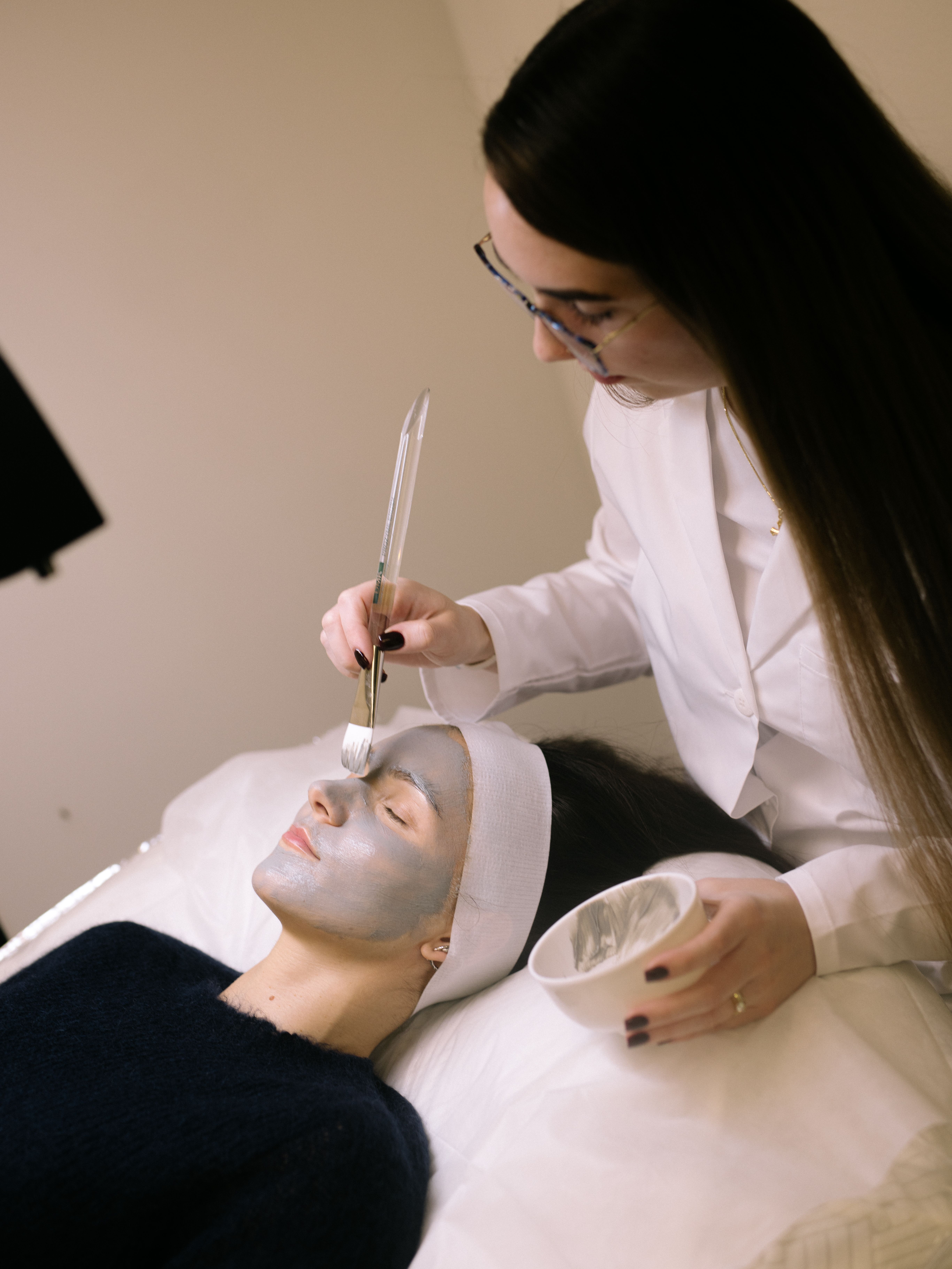 An esthetician applies a grey face mask to a client's face with a brush.