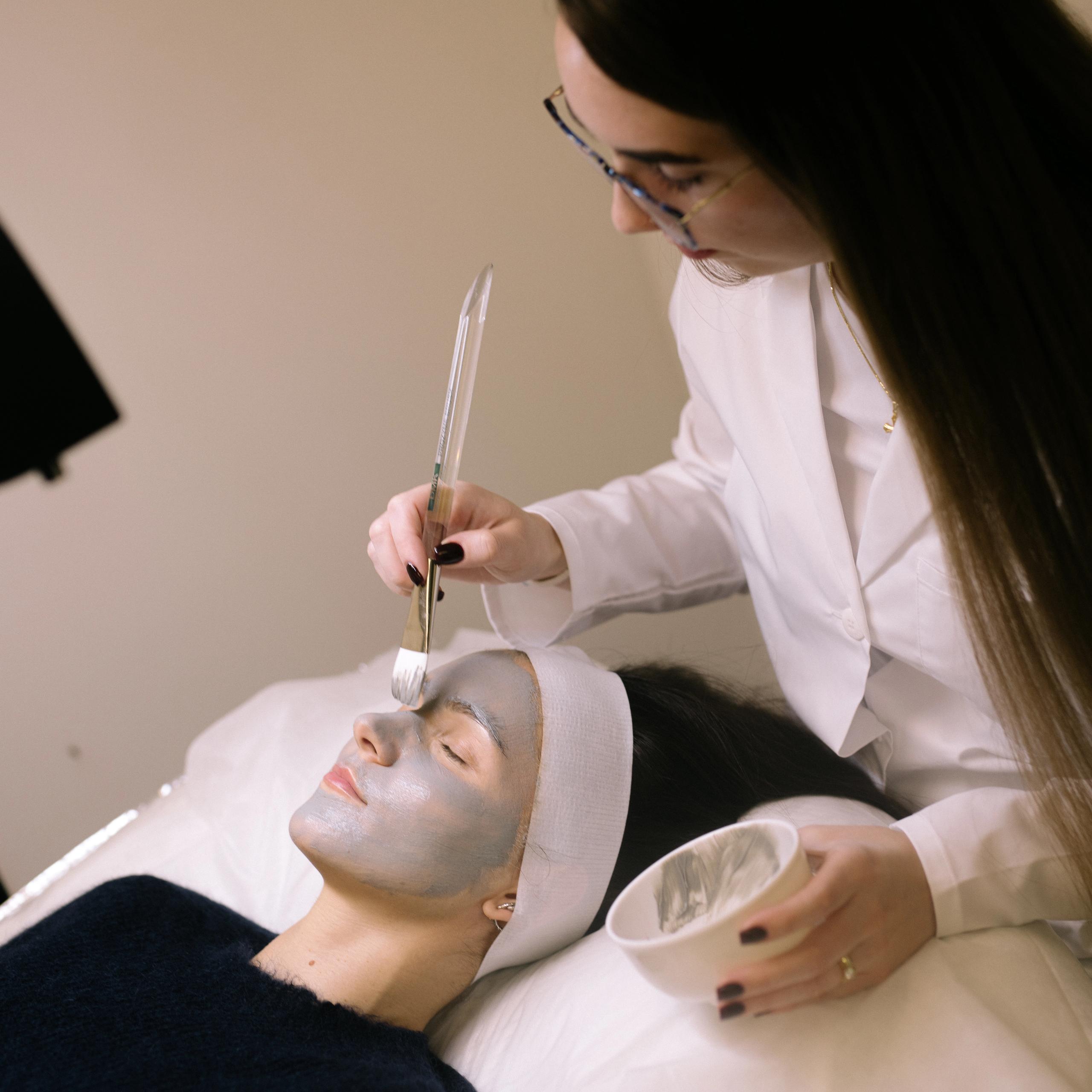 An esthetician applies a grey face mask to a client's face with a brush.