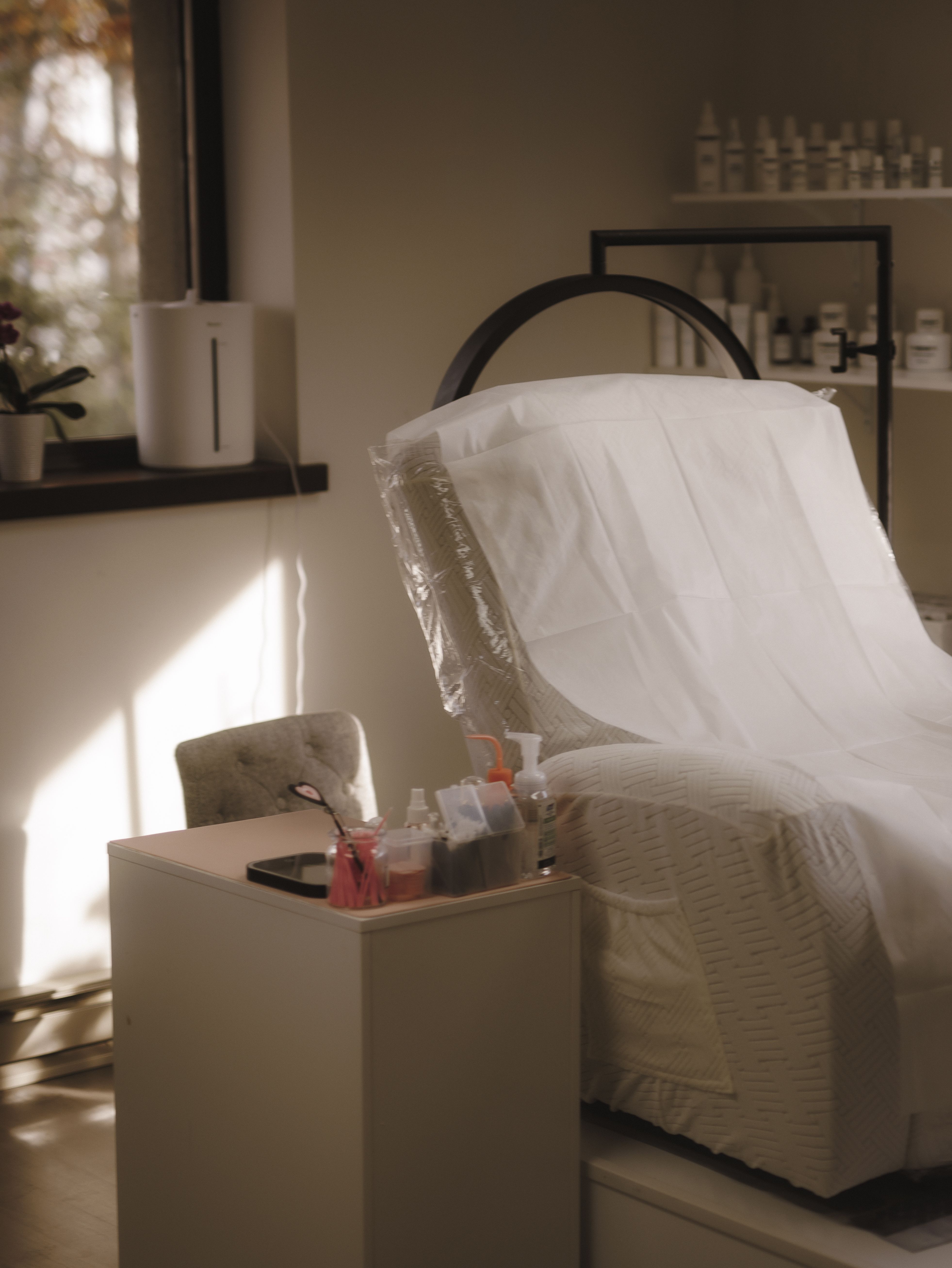 Beauty Republic treatment room with a reclining chair, magnifying lamp, and skincare products beside a sunlit window.