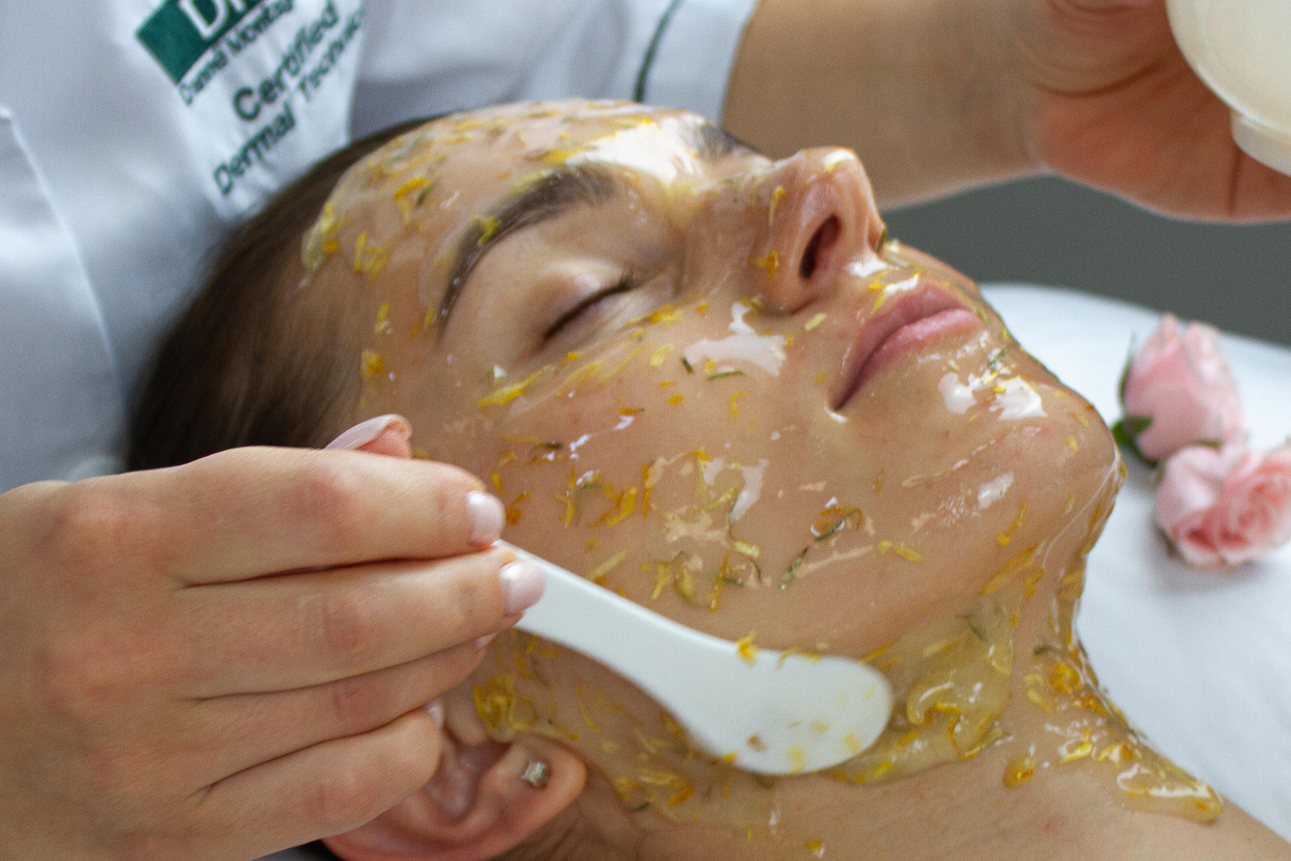 Aesthetician applies a yellow, petal-infused facial mask to a woman's face.