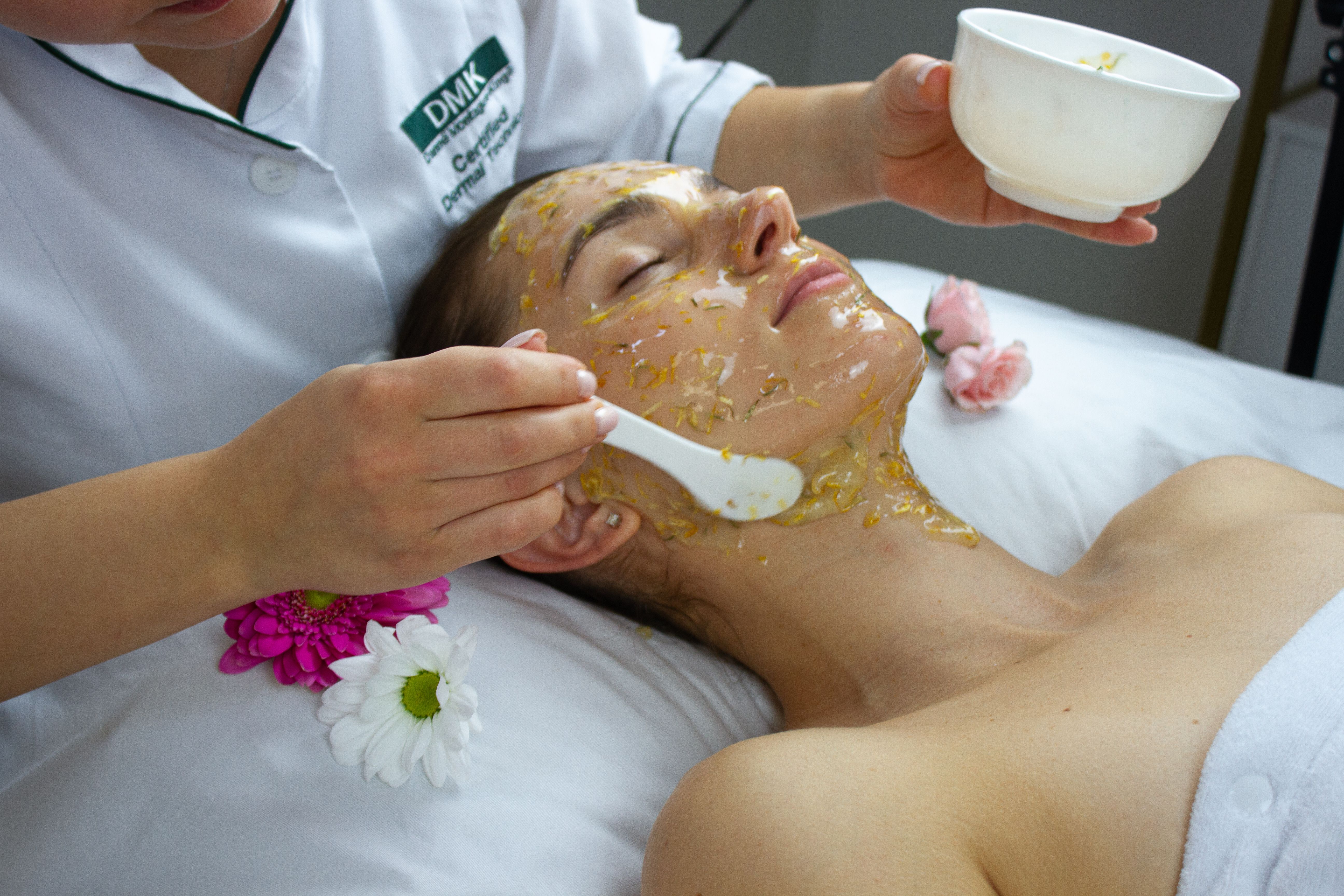 An esthetician applies a floral facial mask to a woman's face and neck in a spa setting.