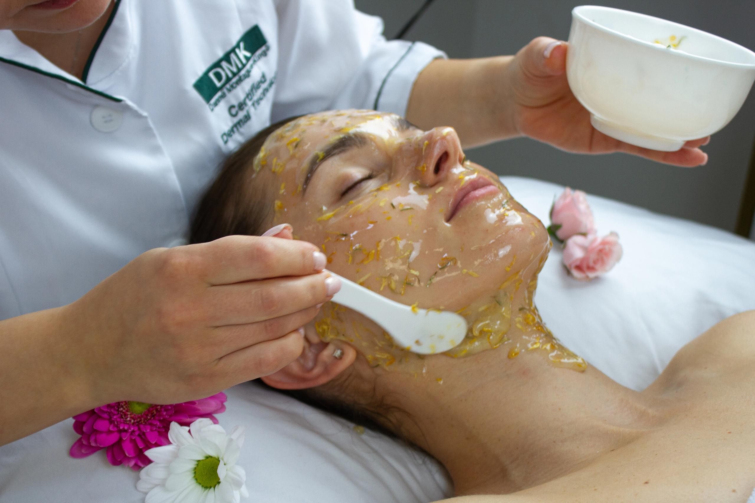 An esthetician applies a floral facial mask to a woman's face and neck in a spa setting.