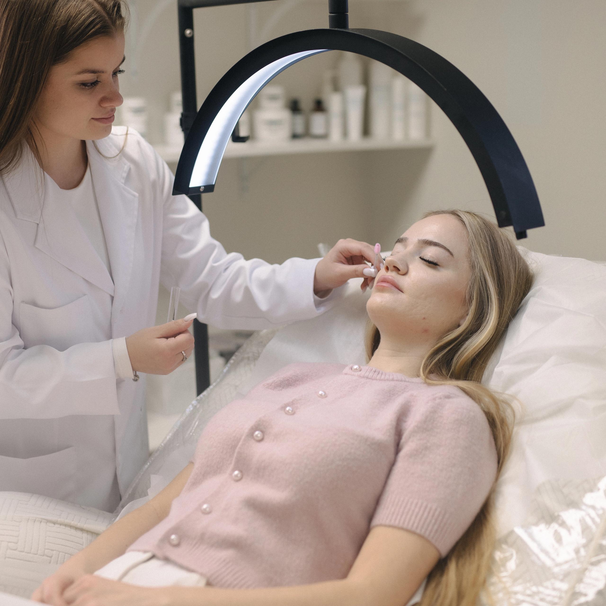 A lash artist brushes client's eyelashes under a curved lamp.
