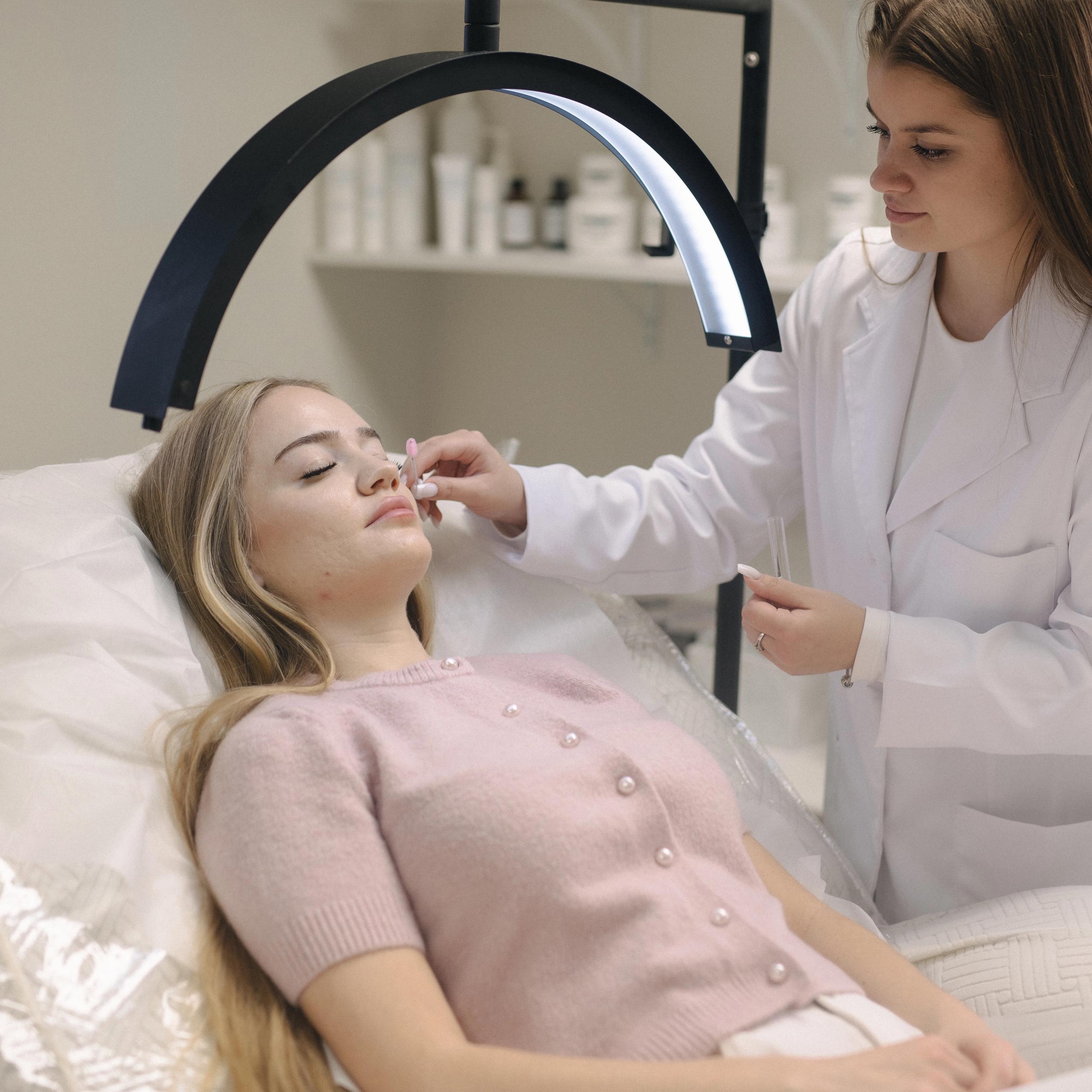 An esthetician brushes client's lashes under a bright ring lamp.