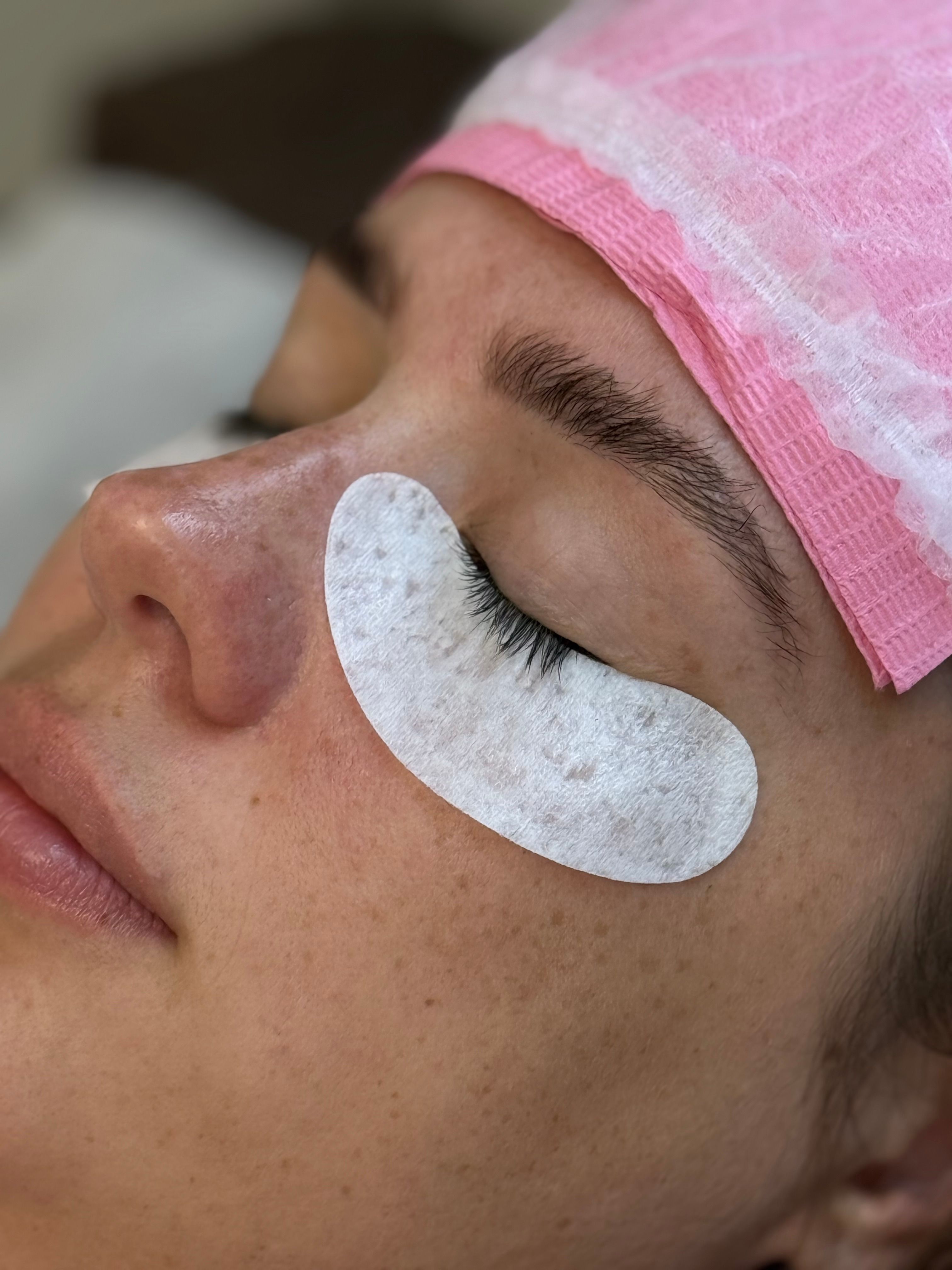 A person with an under-eye pad and pink cap receiving a lash treatment.