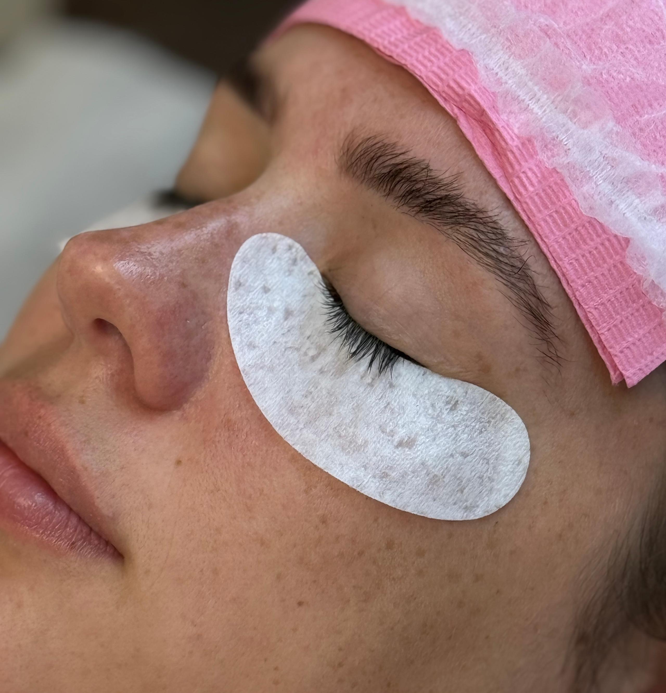A person with an under-eye pad and pink cap receiving a lash treatment.